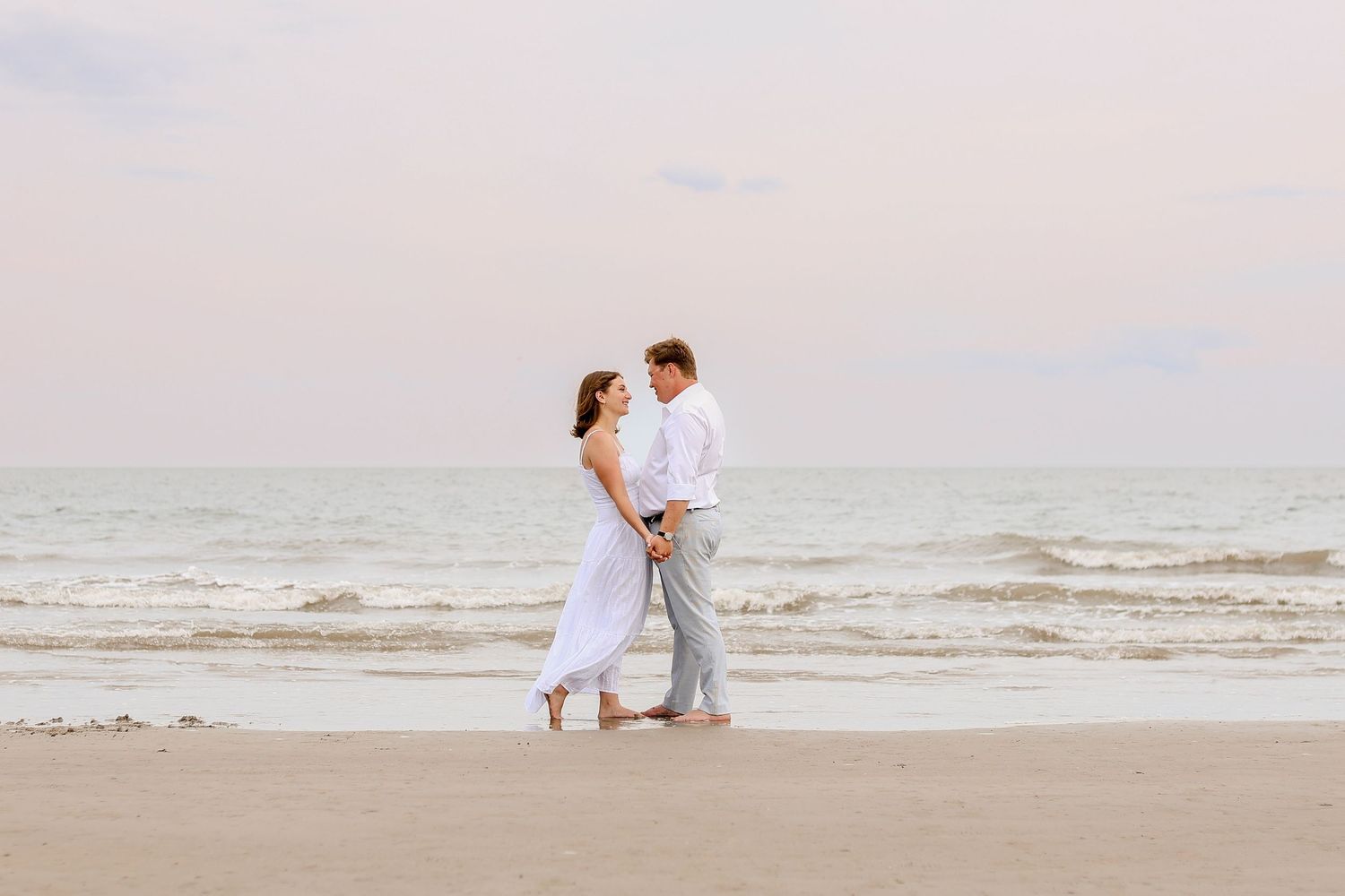 A romantic couple shares tender moments on a serene beach at sunset with waves rolling in the background.