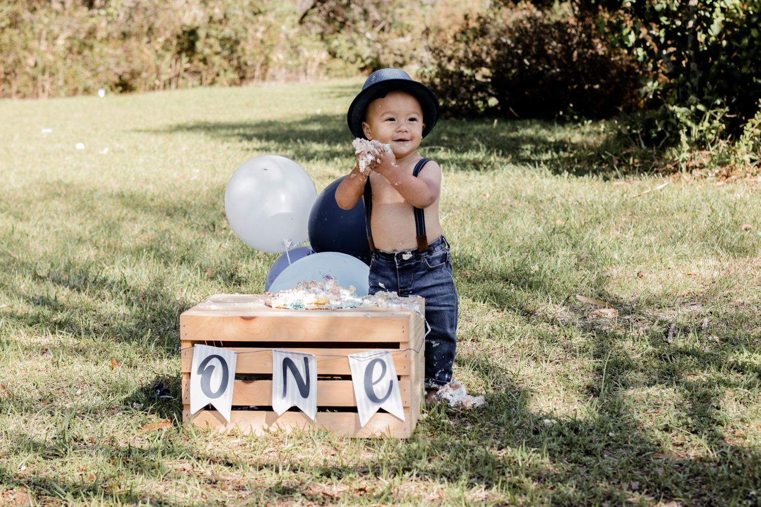 First birthday setup outdoors with blue and white balloons and wooden ONE blocks on grass.