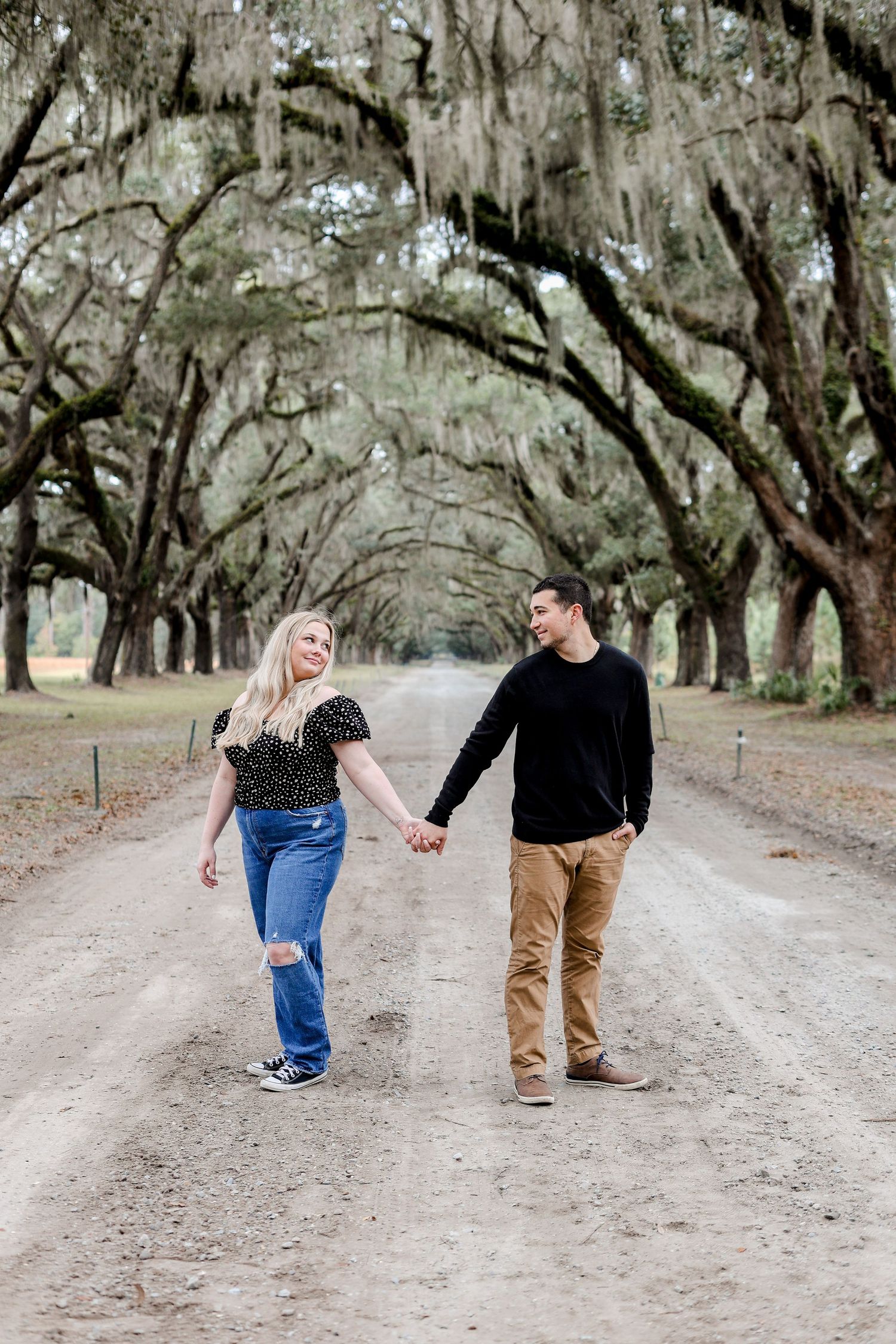 Couple walking hand in hand down a scenic path lined with Spanish moss trees.