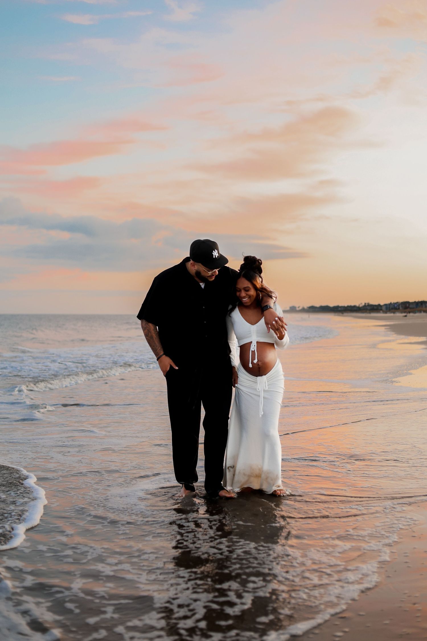 A couple walks along the beach at sunset, their feet in the gentle surf as golden light illuminates the shoreline.