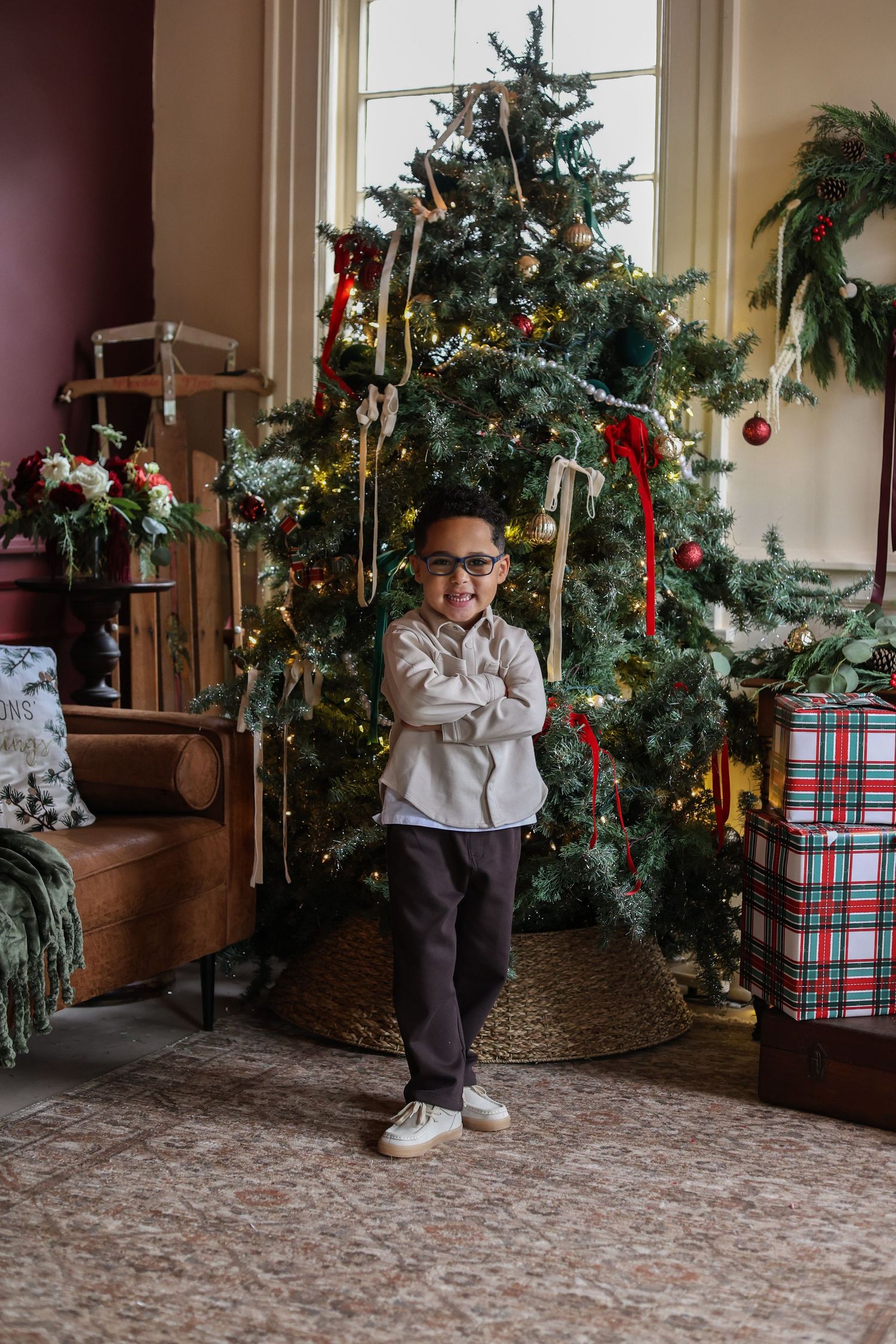 Smiling little boy in glasses and a gray shirt stands with arms crossed beside a decorated Christmas tree and plaid-wrapped gifts.