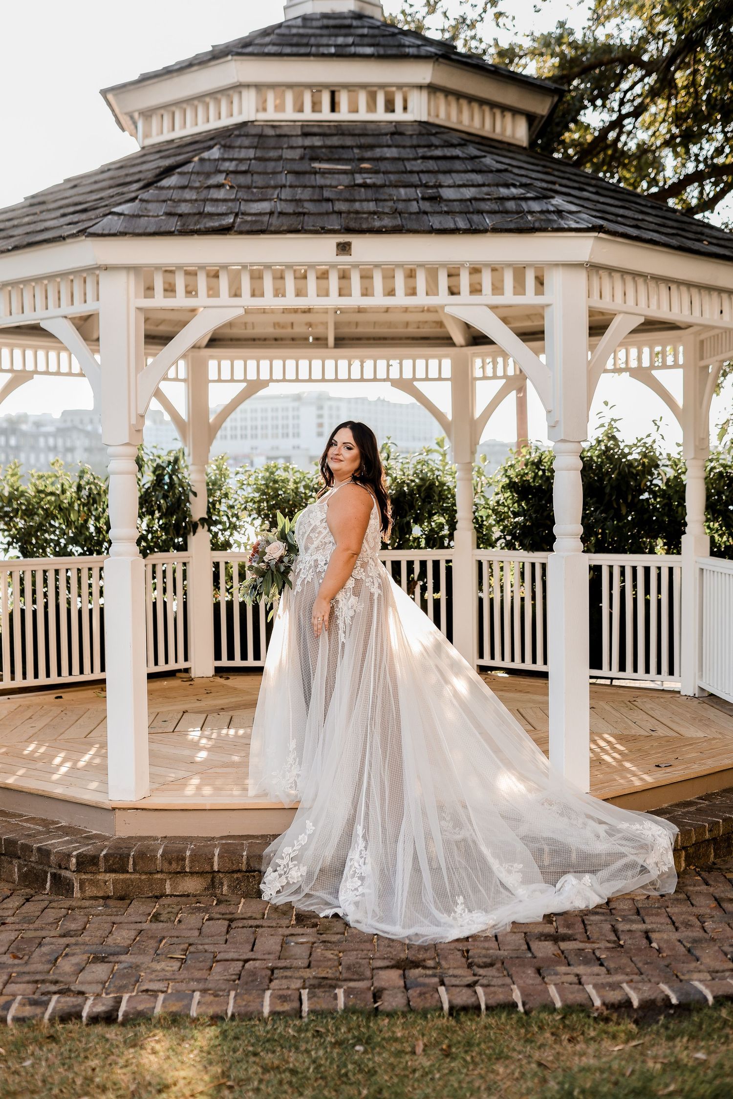 A bride in a flowing white lace gown poses elegantly under a white wooden gazebo during a wedding photoshoot.