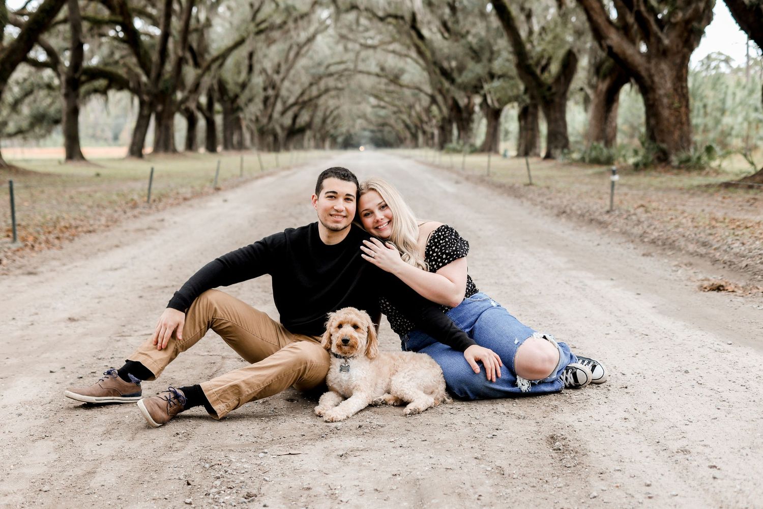 Playful couple lying on a dirt path with their dog during a photo session.