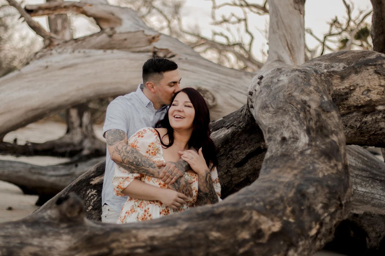 Joyful couple laughing together among driftwood on beach during engagement photo session.