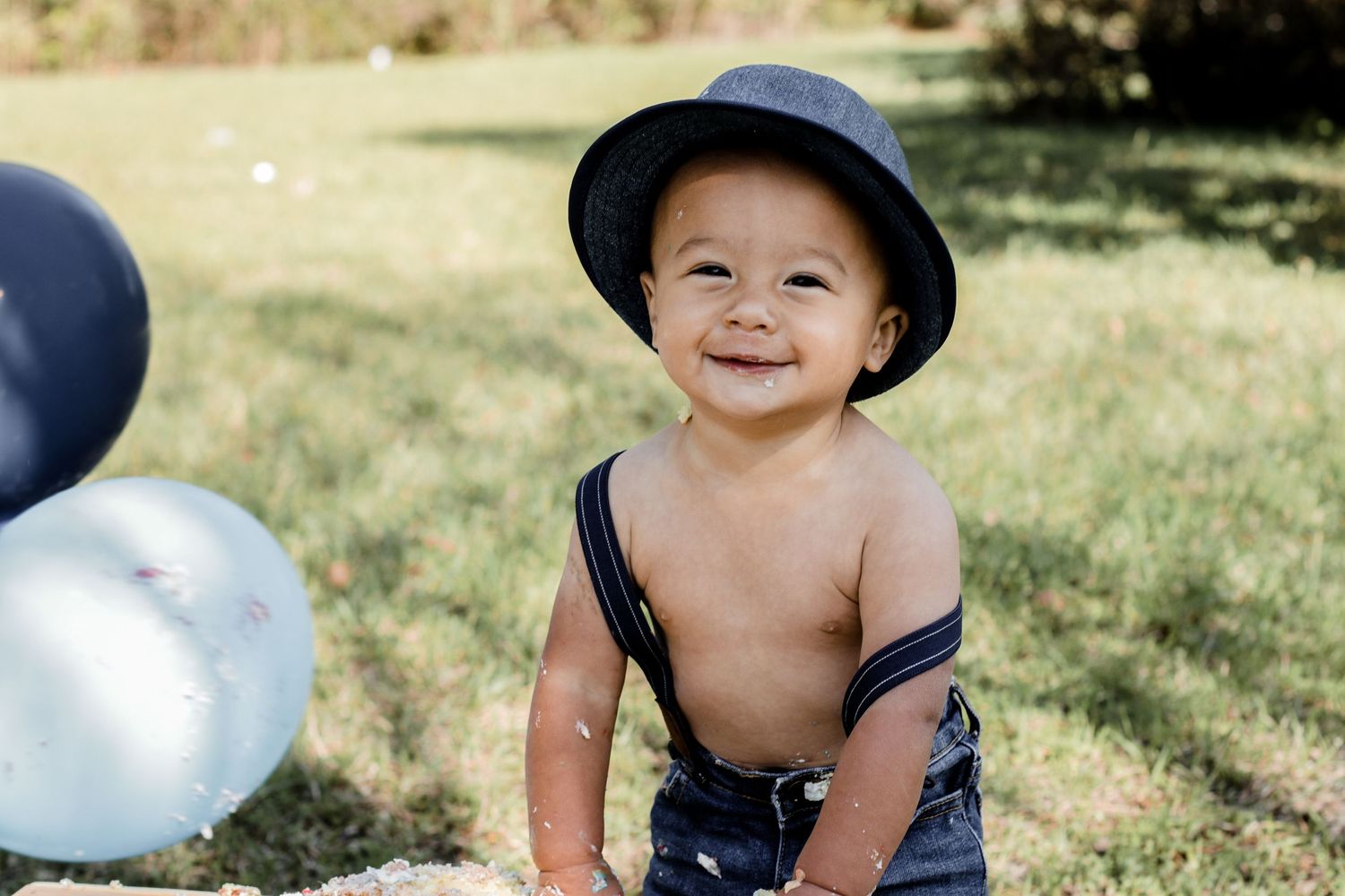 Toddler wearing navy overalls and hat smiles while standing by blue balloons outdoors.