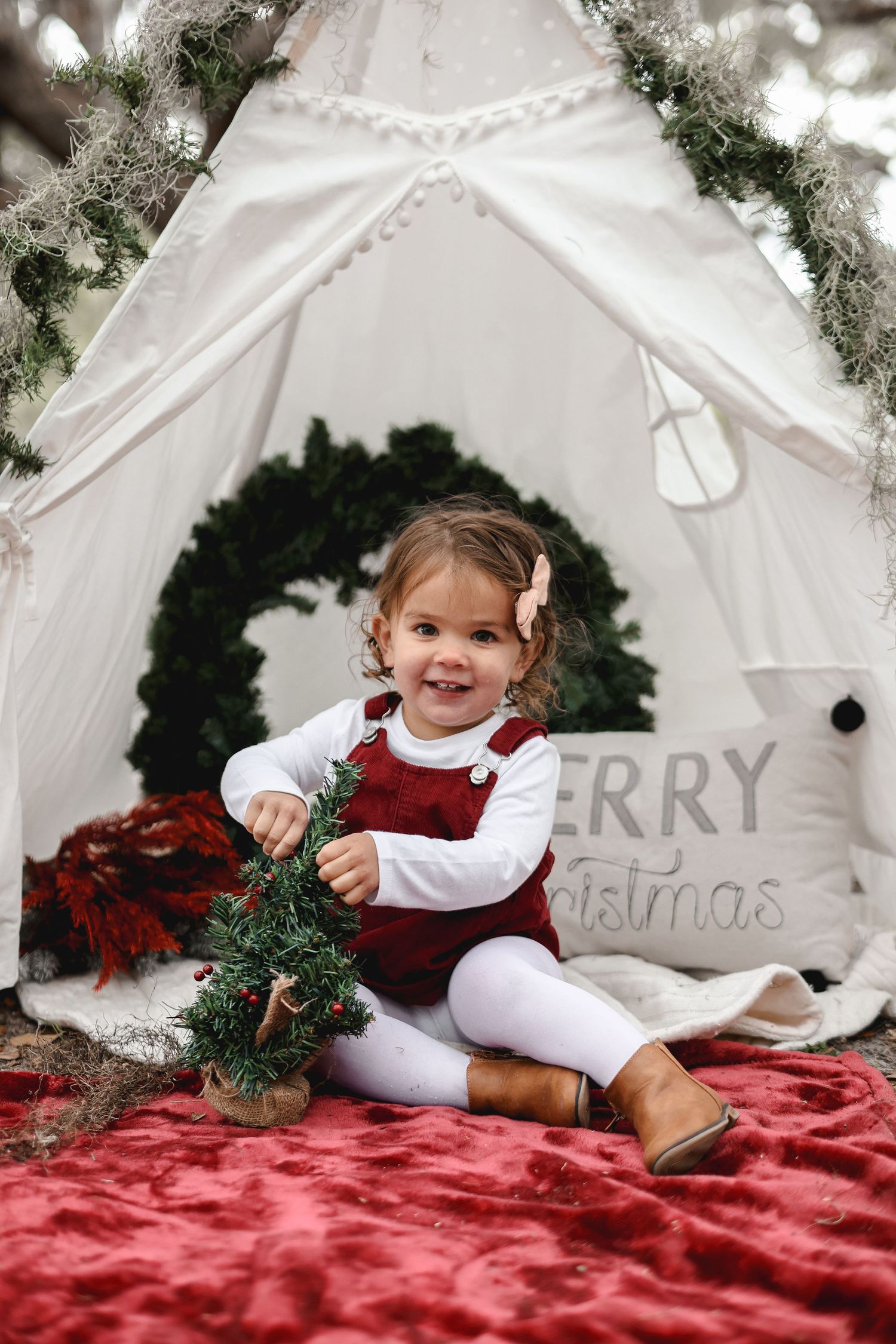A small child in festive attire plays with Christmas decorations inside a white teepee tent.