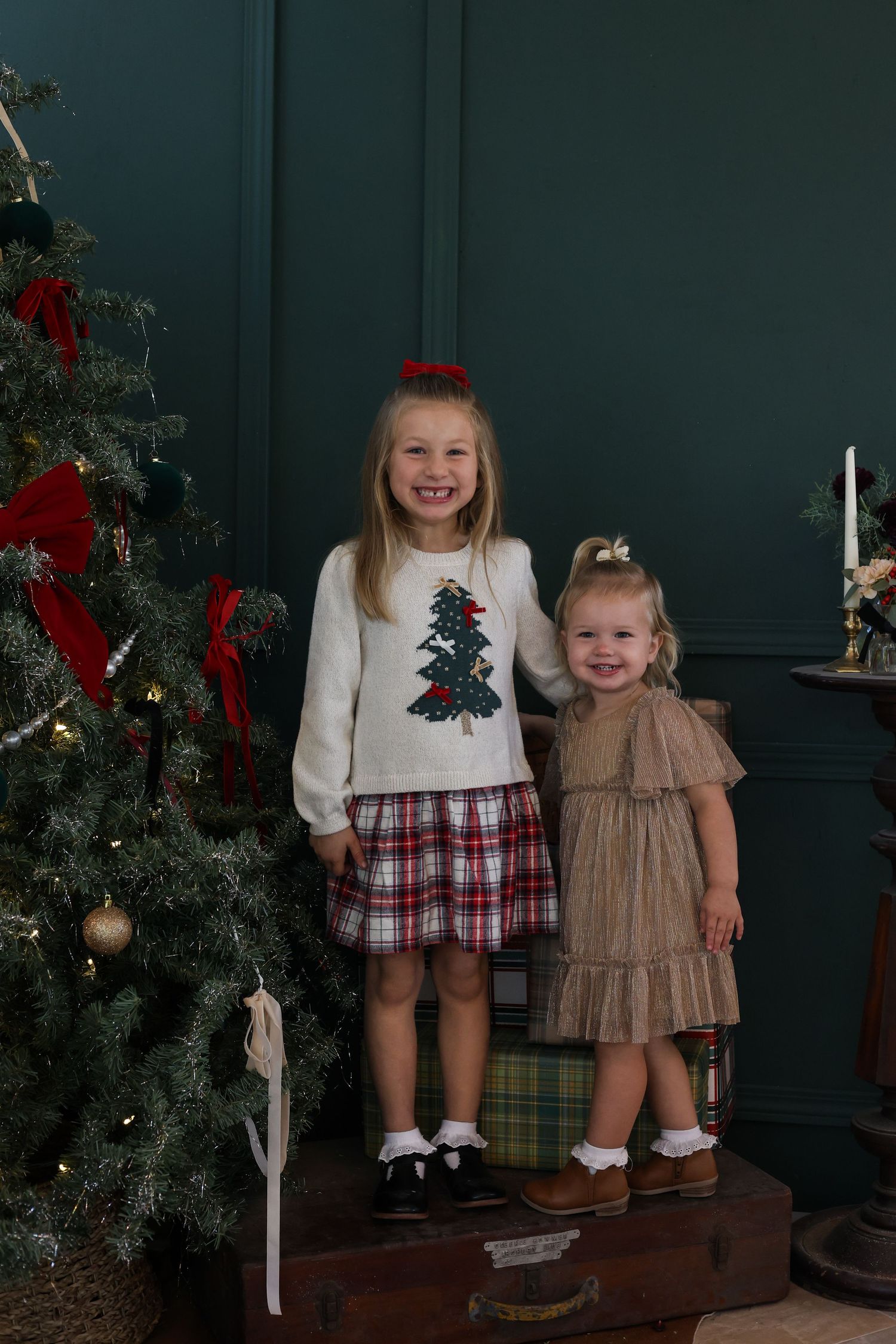 Two children in festive holiday outfits stand together by a decorated Christmas tree with red bows and ornaments.