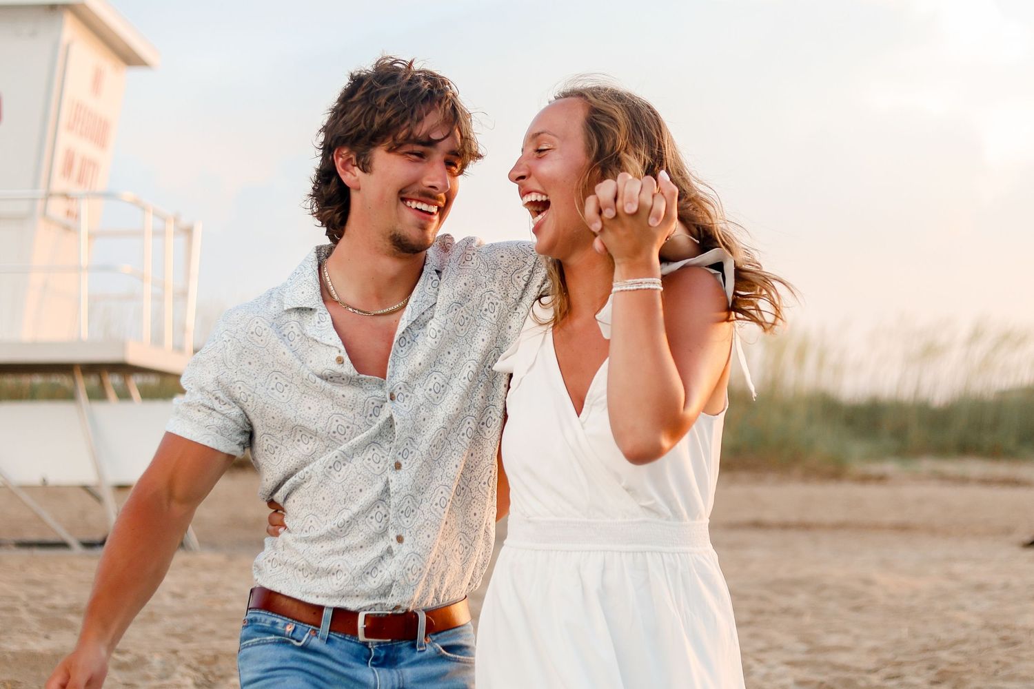 A joyful couple laughing together at sunset on the beach with a lighthouse in the background.