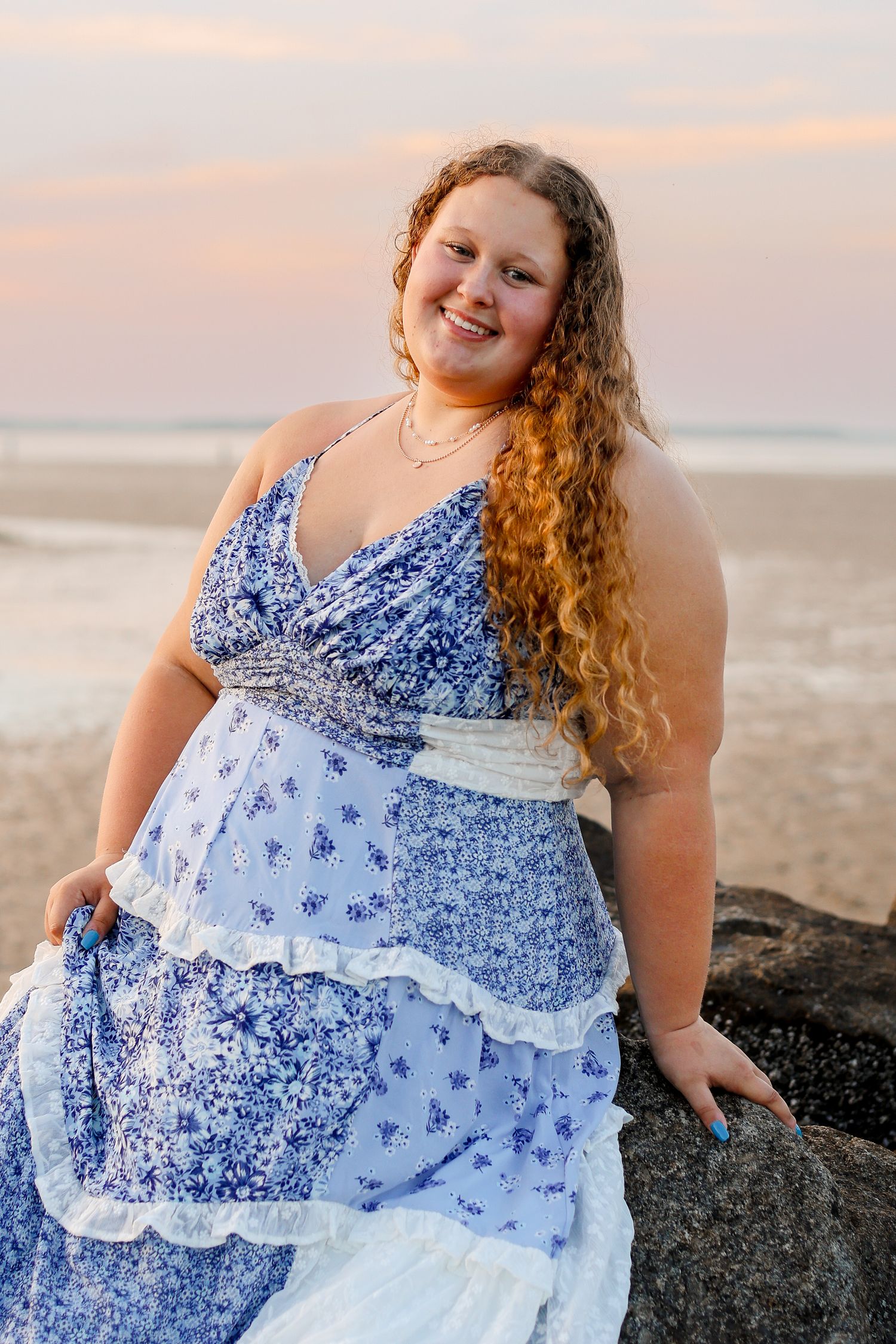 A series of portraits in a blue floral dress sitting on dark rocks at sunset by the ocean.