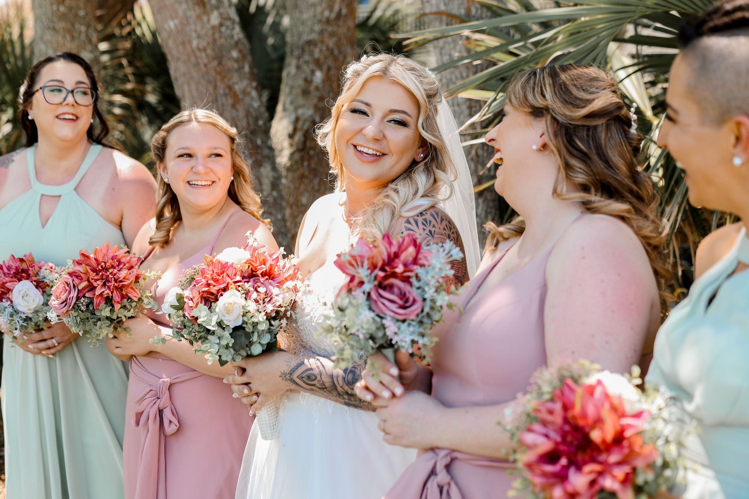 Bridesmaids wearing pastel dresses pose together with colorful bouquets at an outdoor wedding.