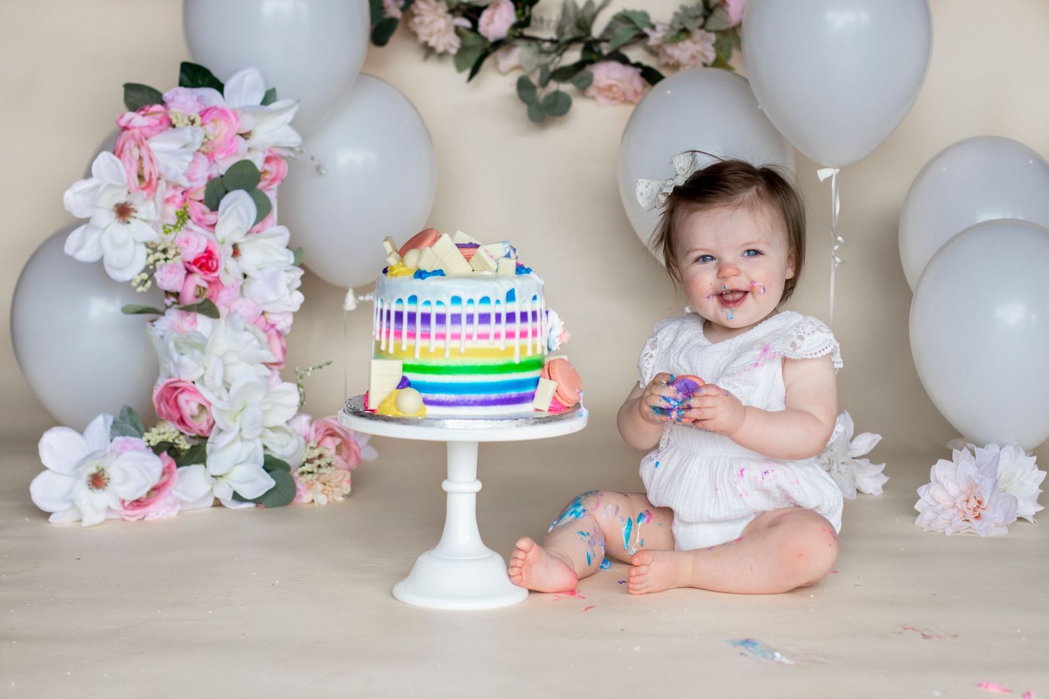 Baby seated in front of white balloons and floral decorations with a small birthday cake.
