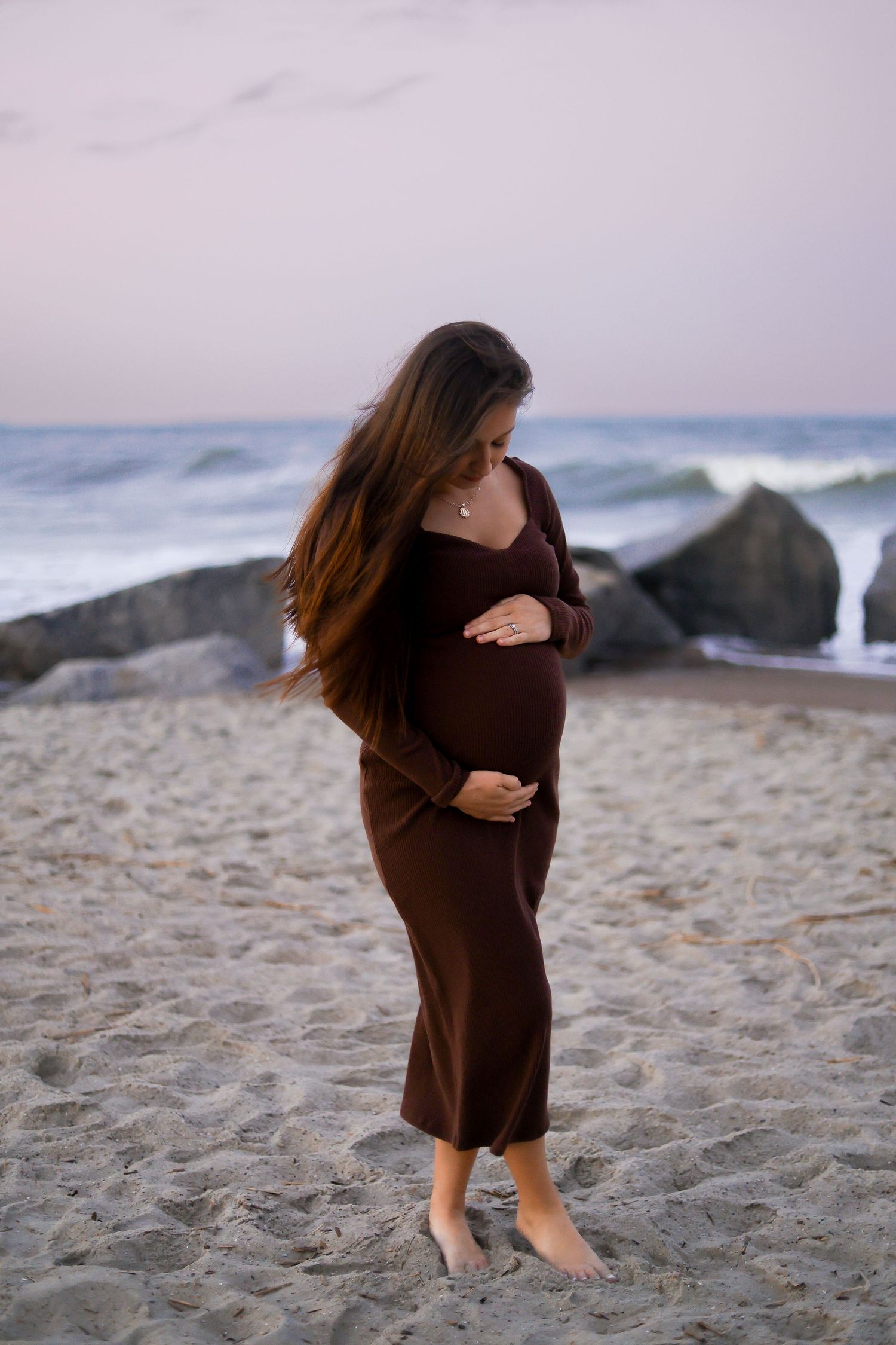 Pregnant woman in brown dress standing barefoot on beach at sunset.