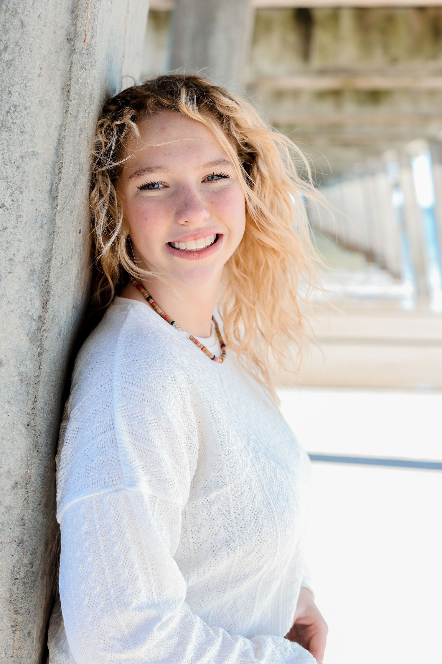 Young woman in white sweater smiling against concrete pillar with soft natural lighting.
