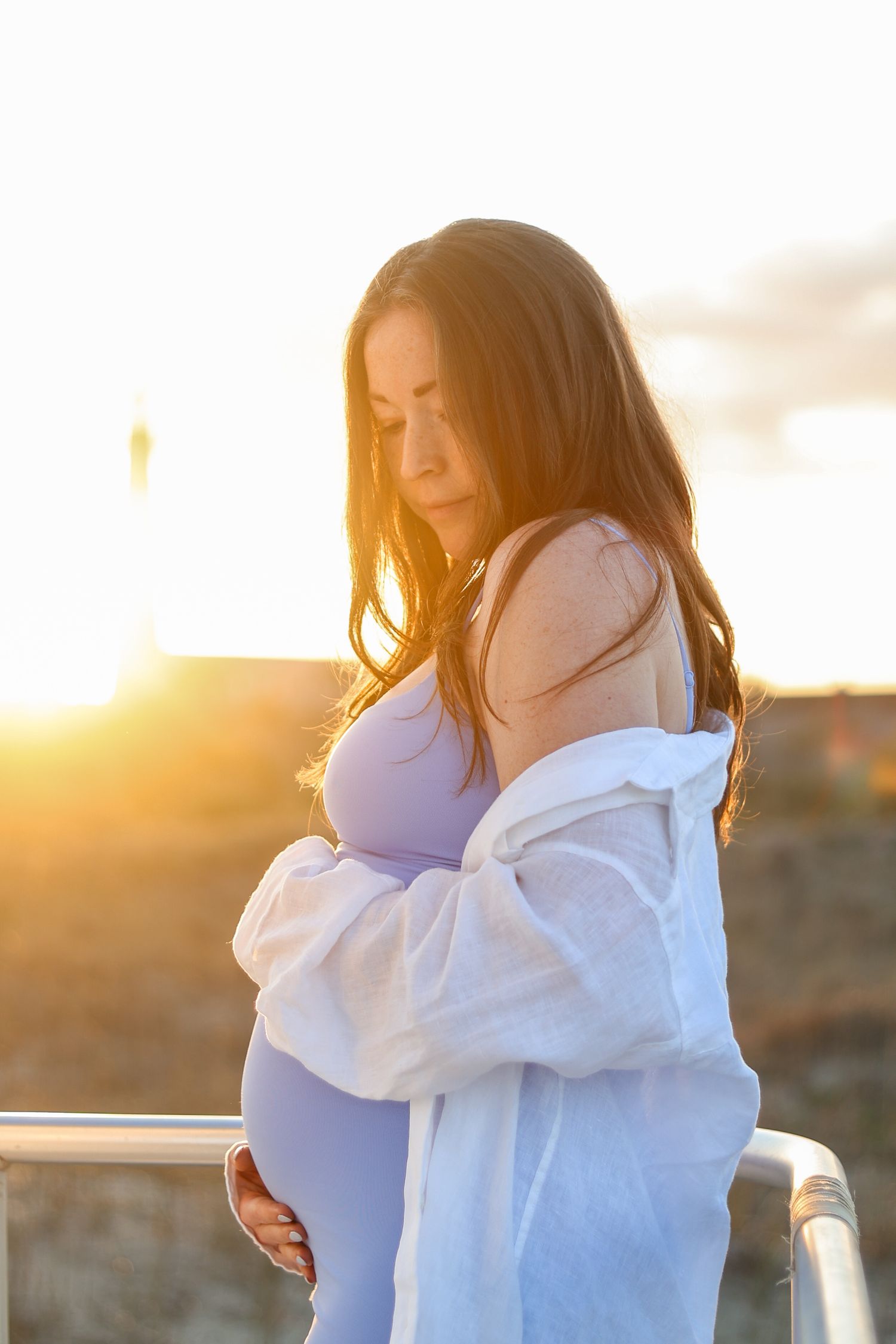 Expectant woman in white dress gazes down at her belly during golden sunset by the beach.