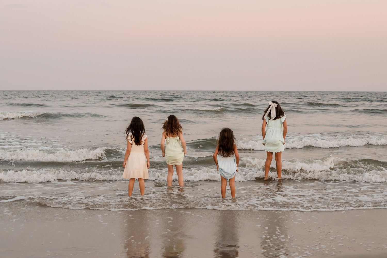 Four people in white dresses stand at the edge of the ocean waves during sunset at the beach.