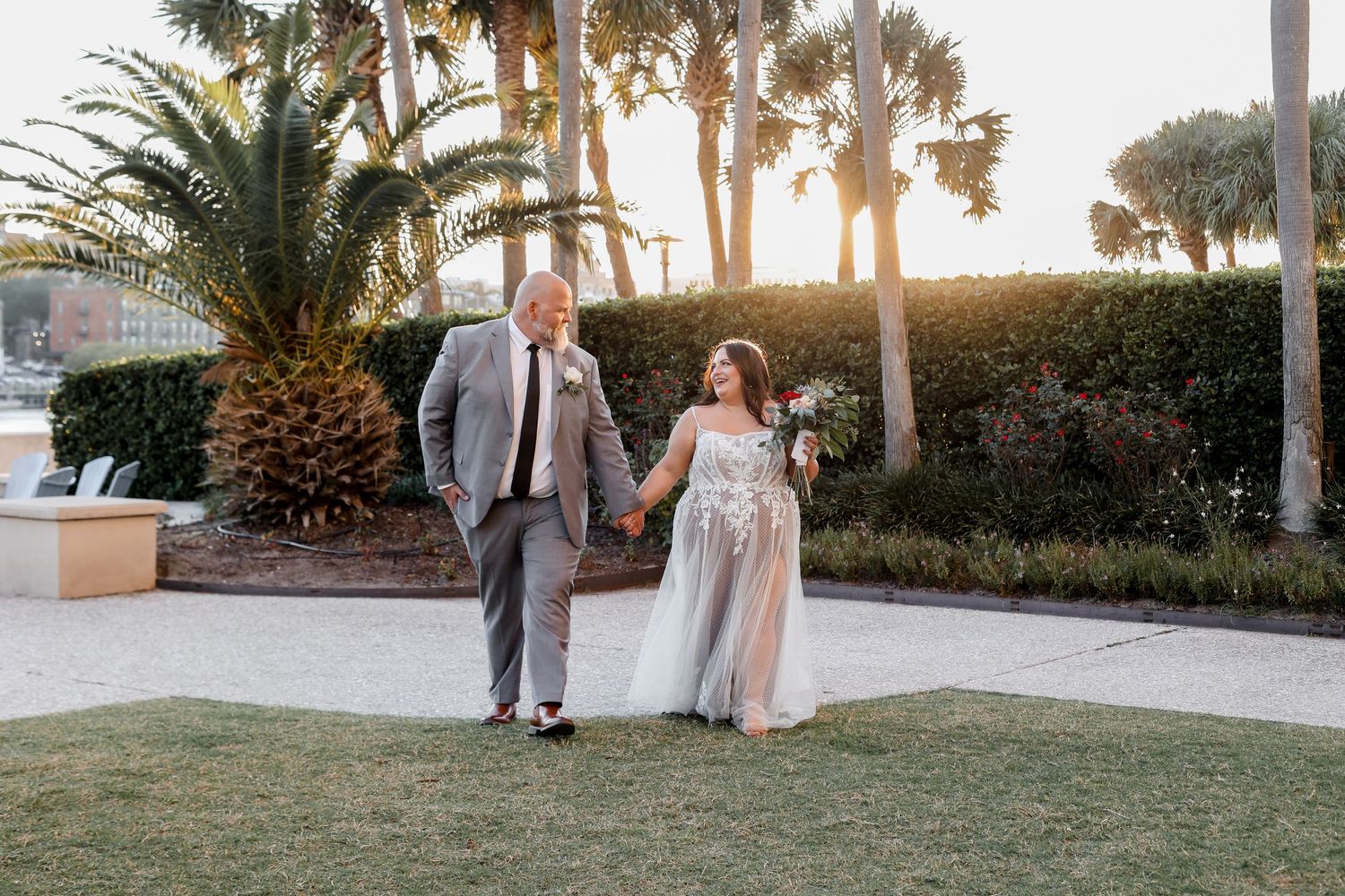 A couple walks hand in hand on a garden path with palm trees and sunset in the background.