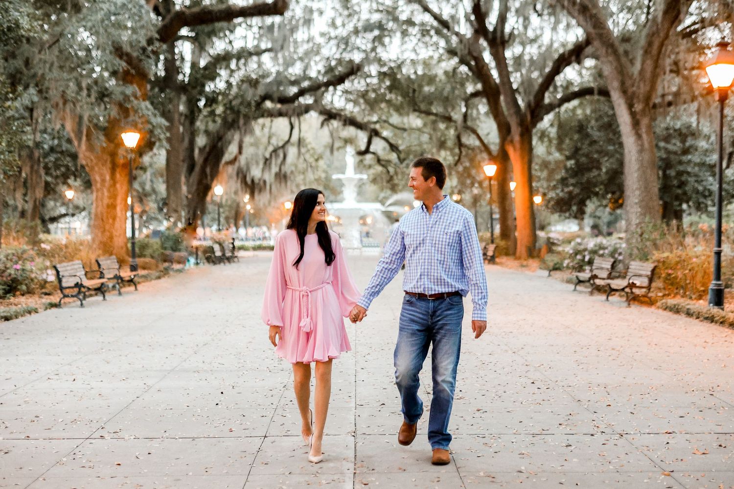 Couple walking hand in hand down tree-lined path at sunset in romantic park setting.