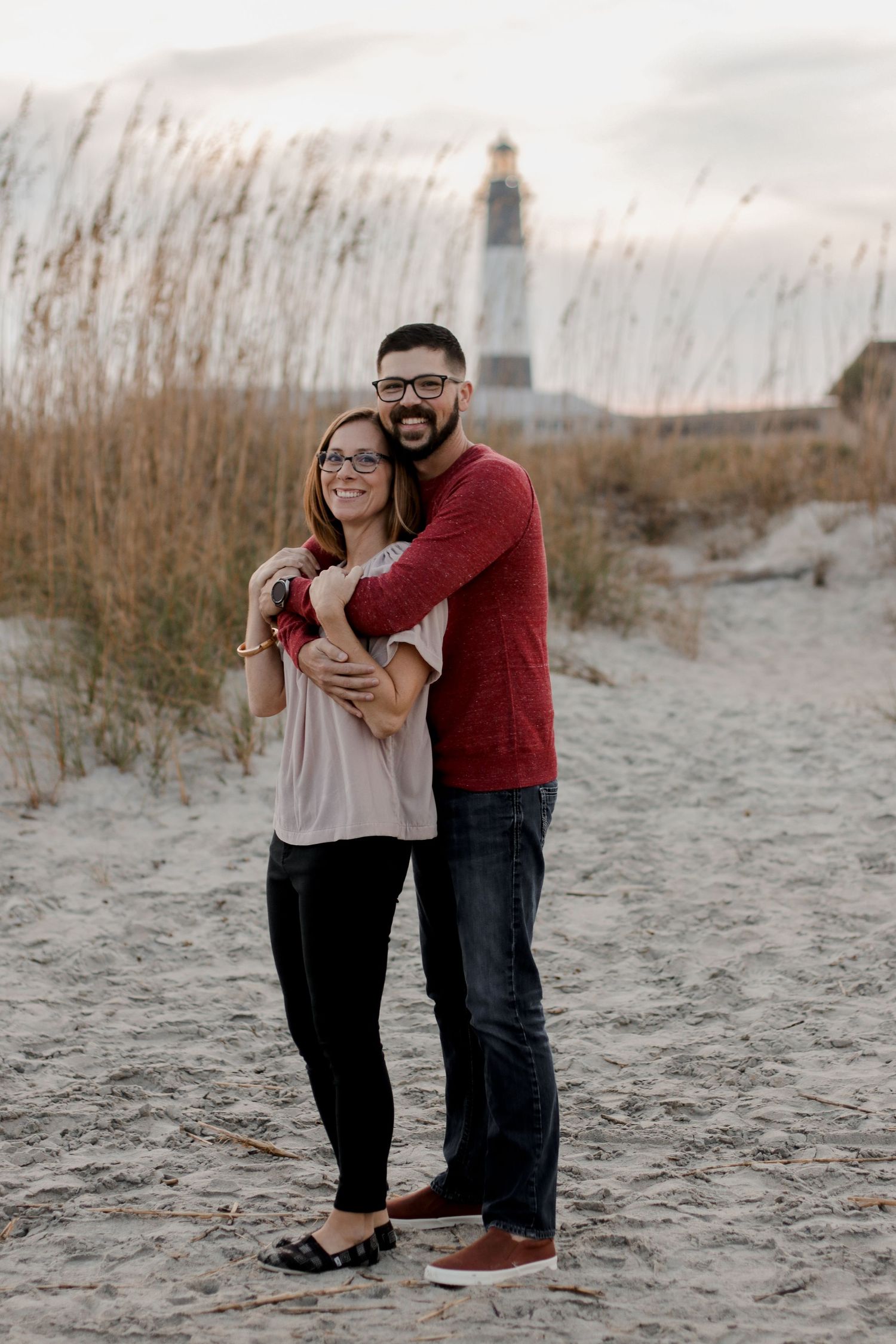 A couple poses among tall beach grass with a lighthouse visible in the background.