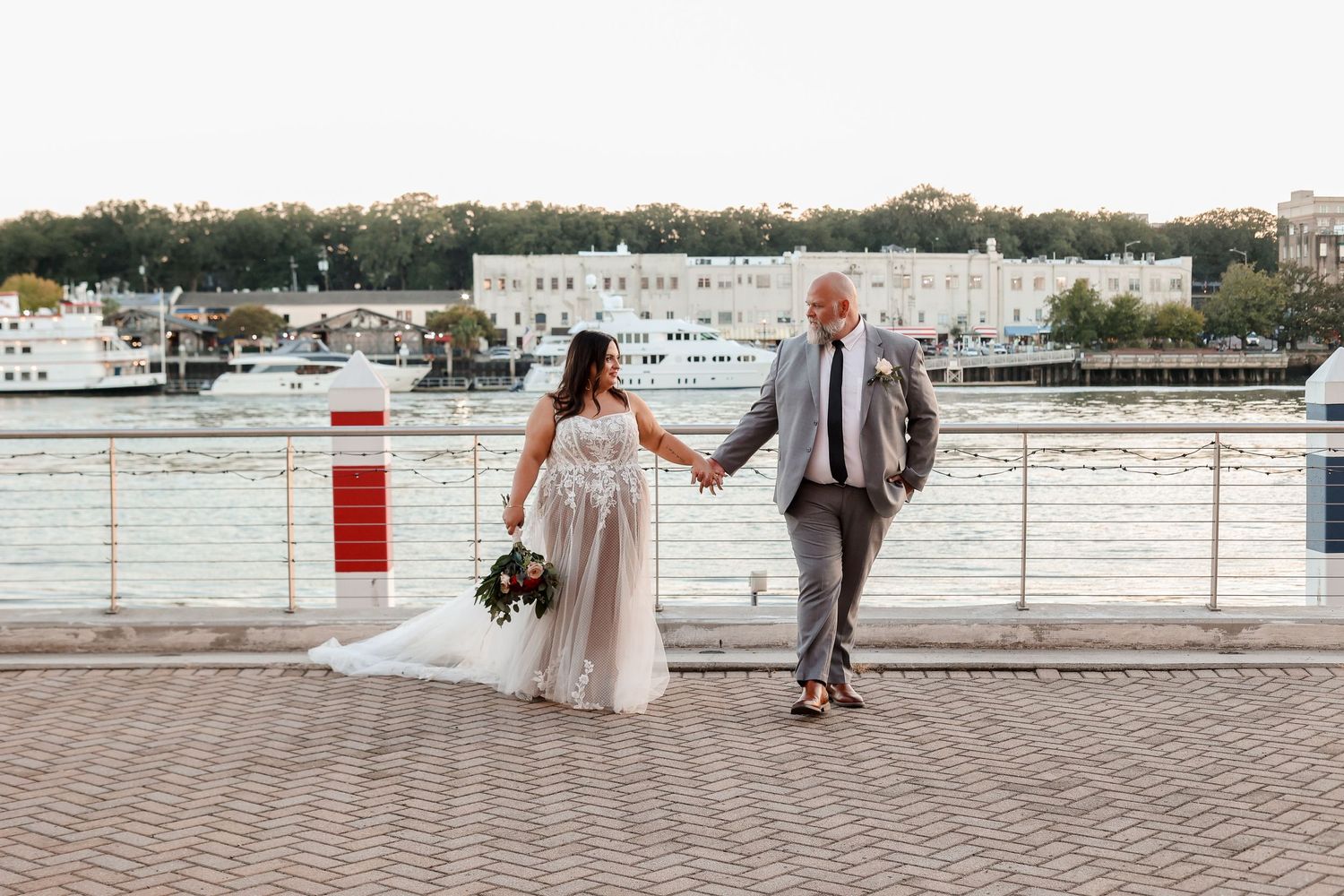 A couple shares a candid moment while walking along a marina boardwalk in their wedding attire.