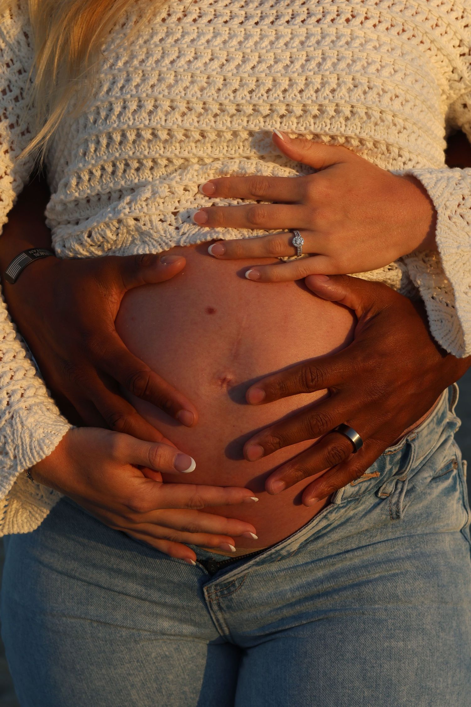 Close-up shots of hands cradling a pregnant belly while wearing a white knit sweater and jeans.