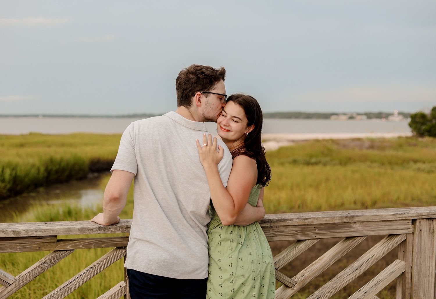 A romantic couple embraces while standing on a wooden boardwalk overlooking coastal marshland at sunset.