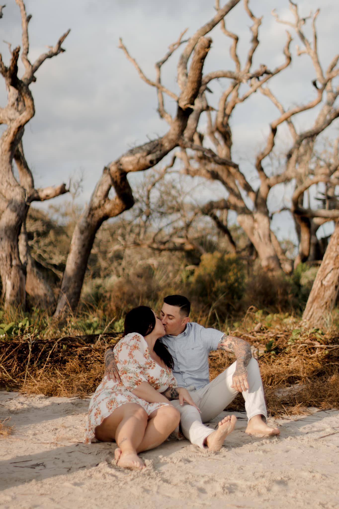 Couple sharing intimate moment on beach surrounded by weathered tree branches at sunset.