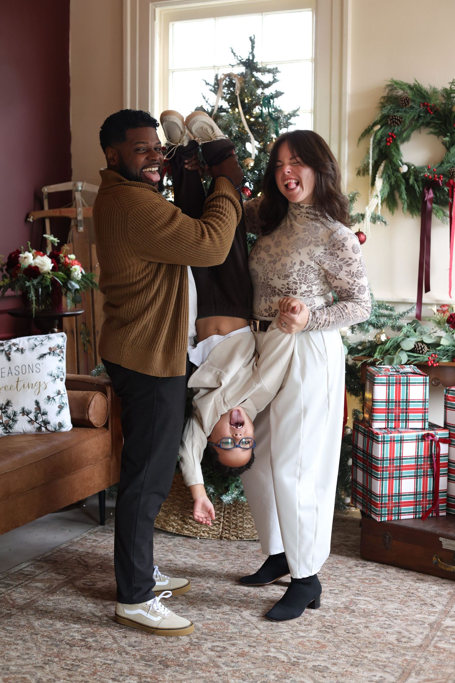 Two people laugh in a festive living room as one playfully holds the other upside down near a Christmas tree, wreath, and wrapped gifts.