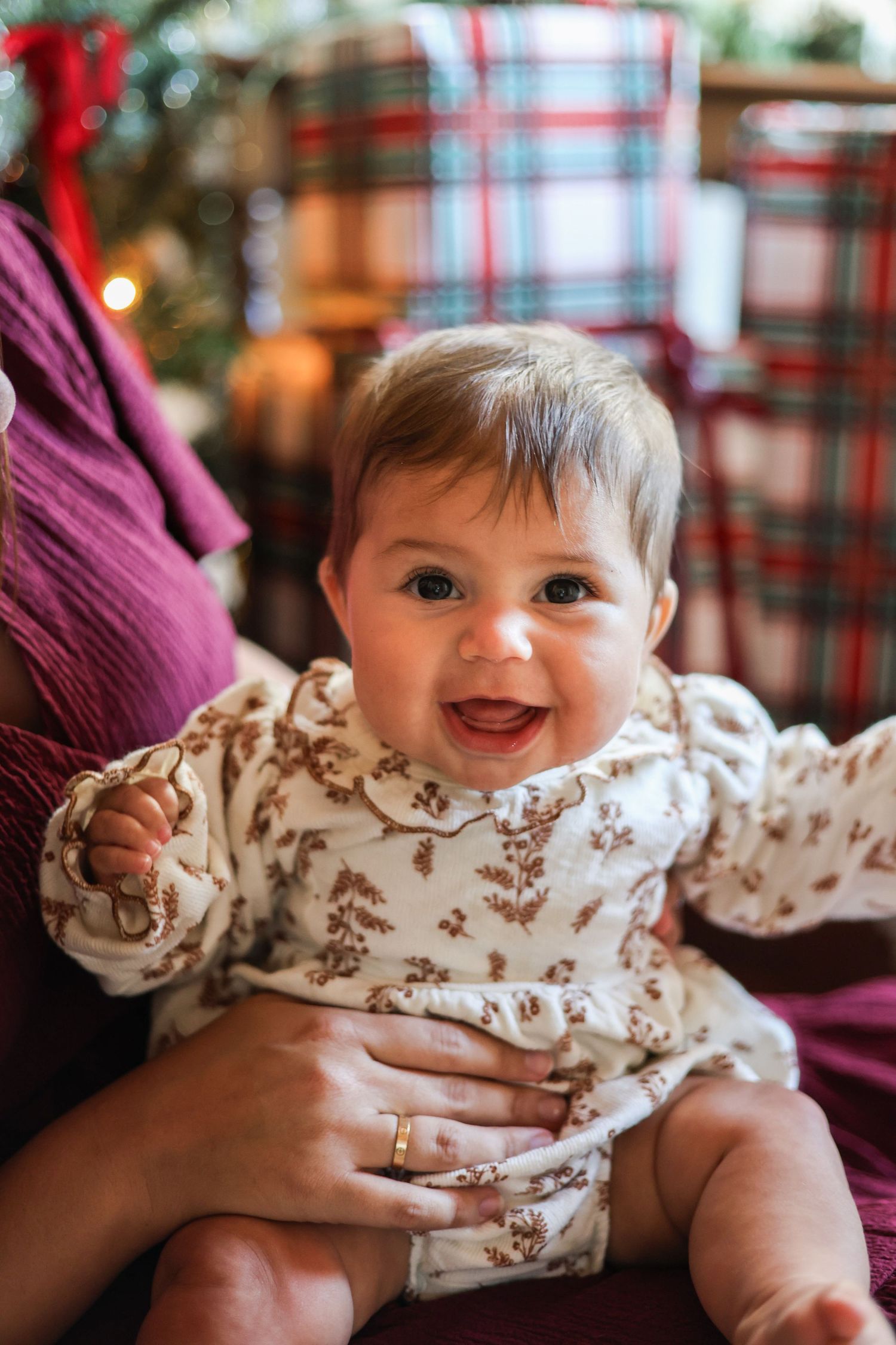 Smiling baby in a floral cream dress sits on an adult's lap in front of plaid wrapped Christmas gifts and a glowing tree.
