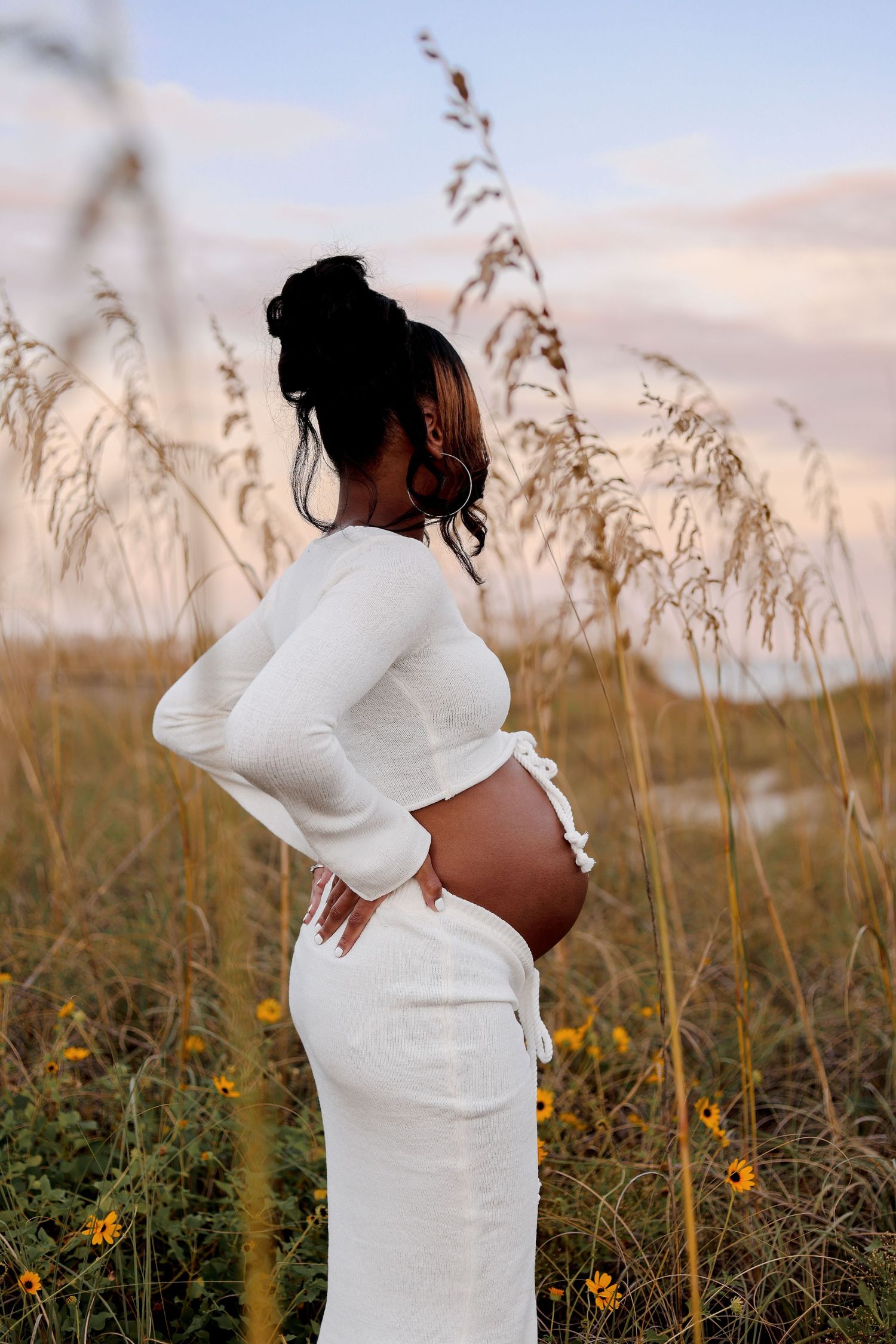 Silhouette of a person in a white dress poses against tall grass and wildflowers during golden hour.