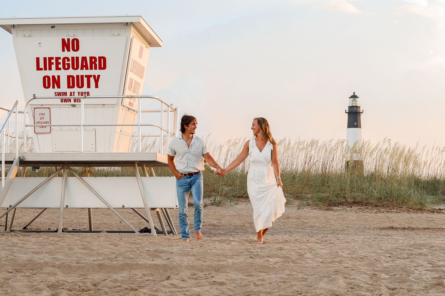 A couple holds hands walking on a beach by a lifeguard tower with a lighthouse visible in the distance at dusk.