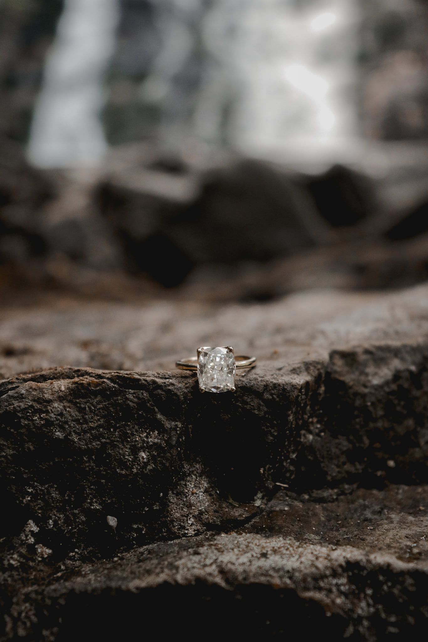 Diamond engagement ring sitting on dark textured rock surface with blurred waterfall background.