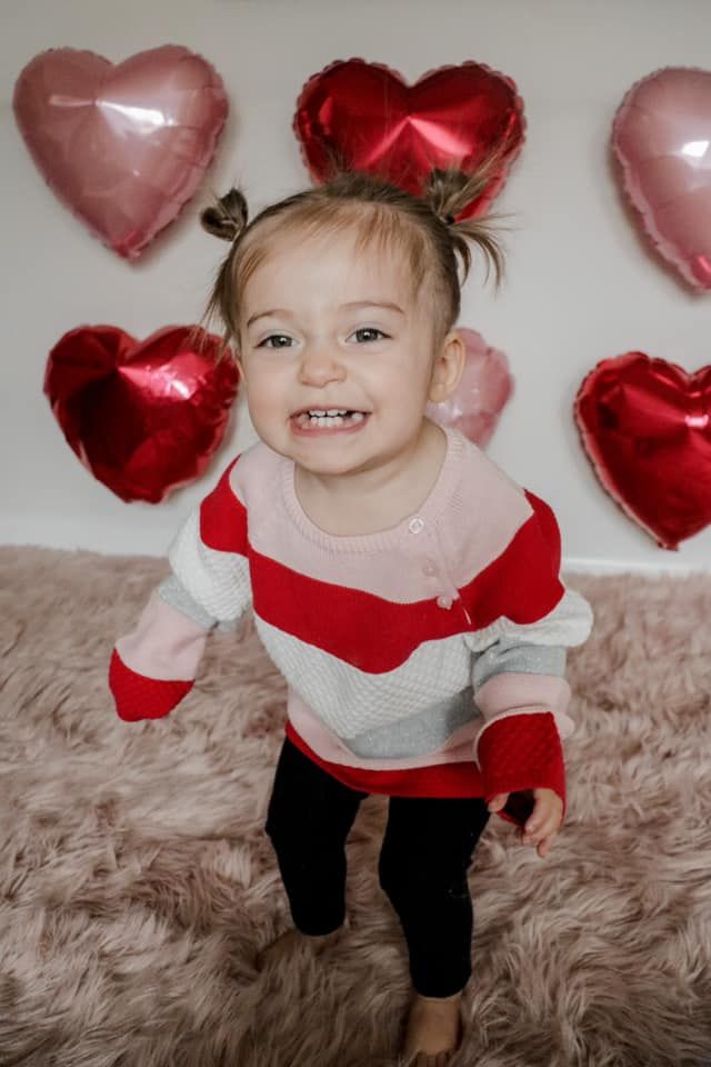 Toddler in red and white outfit standing on furry rug with heart balloons on wall.