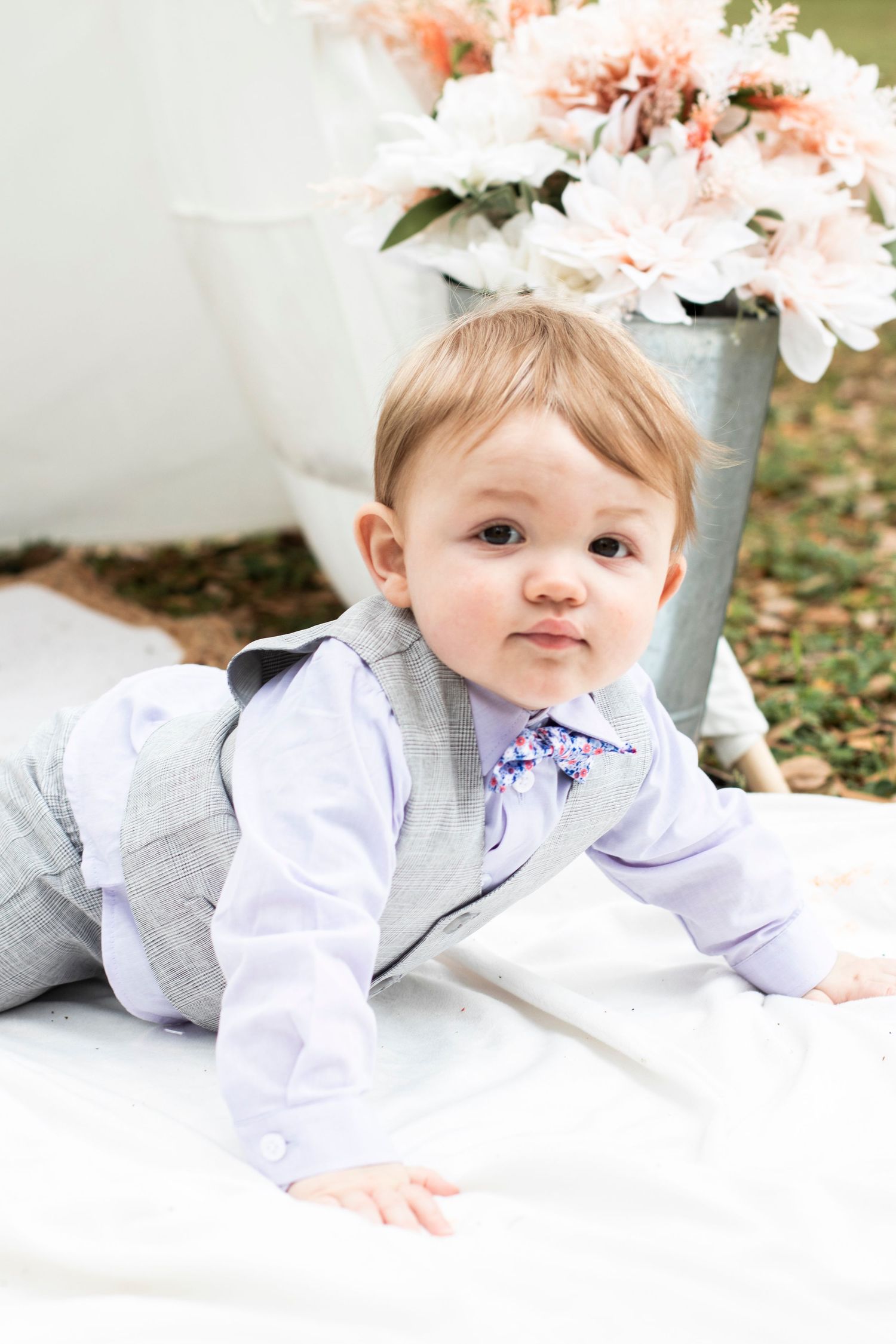 Baby in gray and white outfit lying on white blanket surrounded by soft pink floral decor.