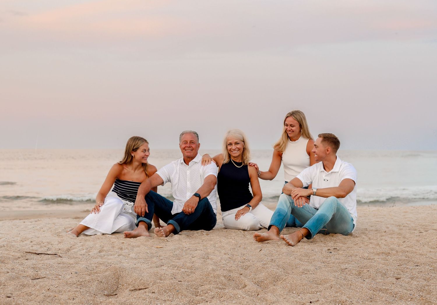 A family group sits together on a sandy beach at sunset, laughing and enjoying the peaceful coastal setting.