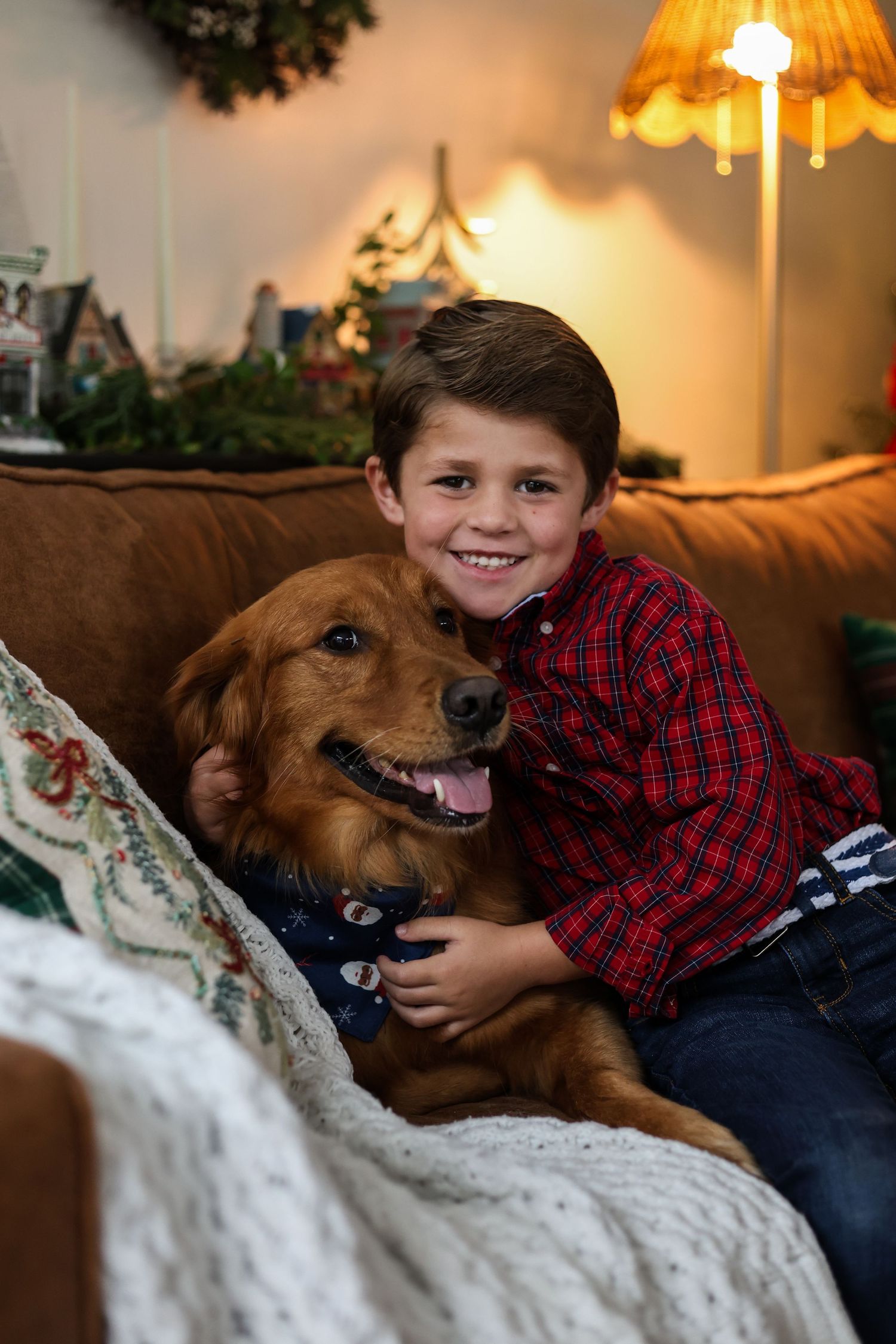 A child in a red plaid shirt cuddles with a brown dog on a white couch in a warmly lit living room.