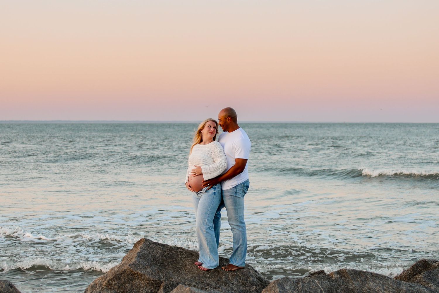 A couple embraces on rocky beach during sunset with ocean waves crashing in the background.