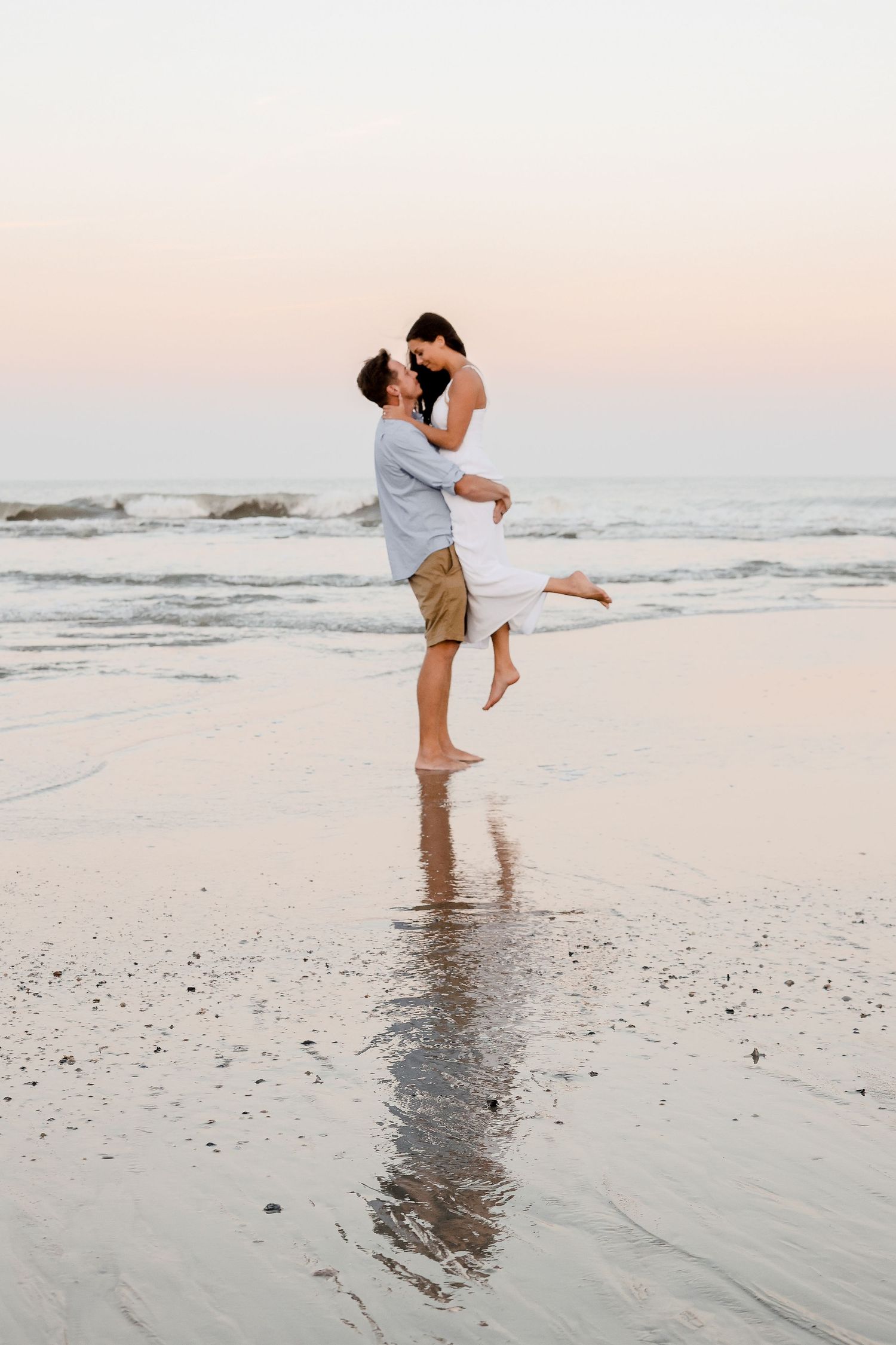 A joyful couple embraces on the beach with one person lifted in the air against the sunset.
