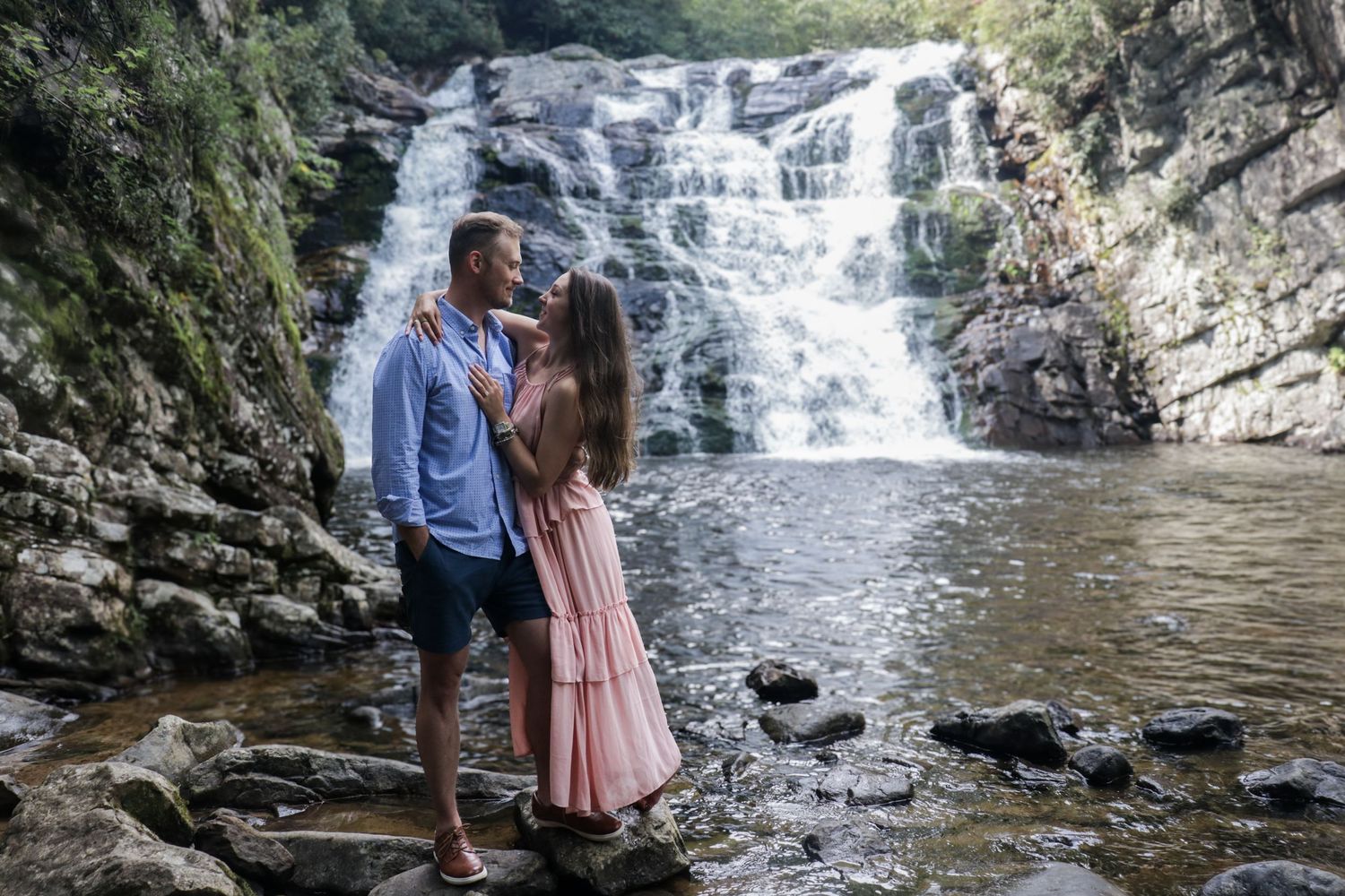 A couple in casual attire shares an intimate moment in front of a scenic waterfall.