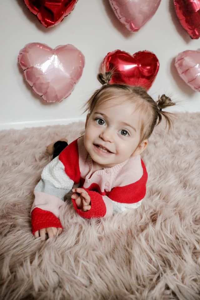 A baby in red and white holiday outfit smiles beneath heart-shaped balloons.