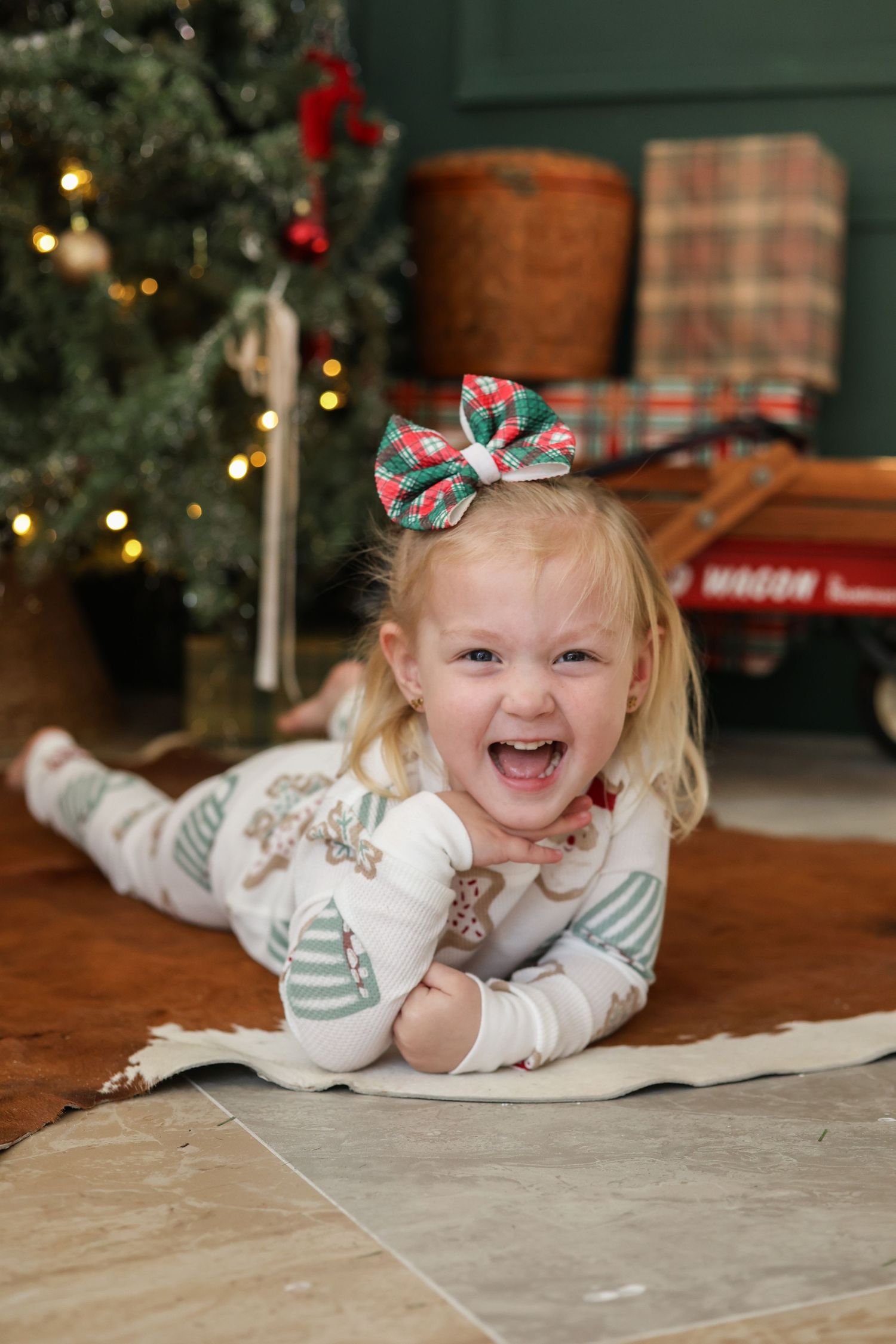 A child wearing a holiday bow and white outfit lies on the floor smiling near a decorated Christmas tree.