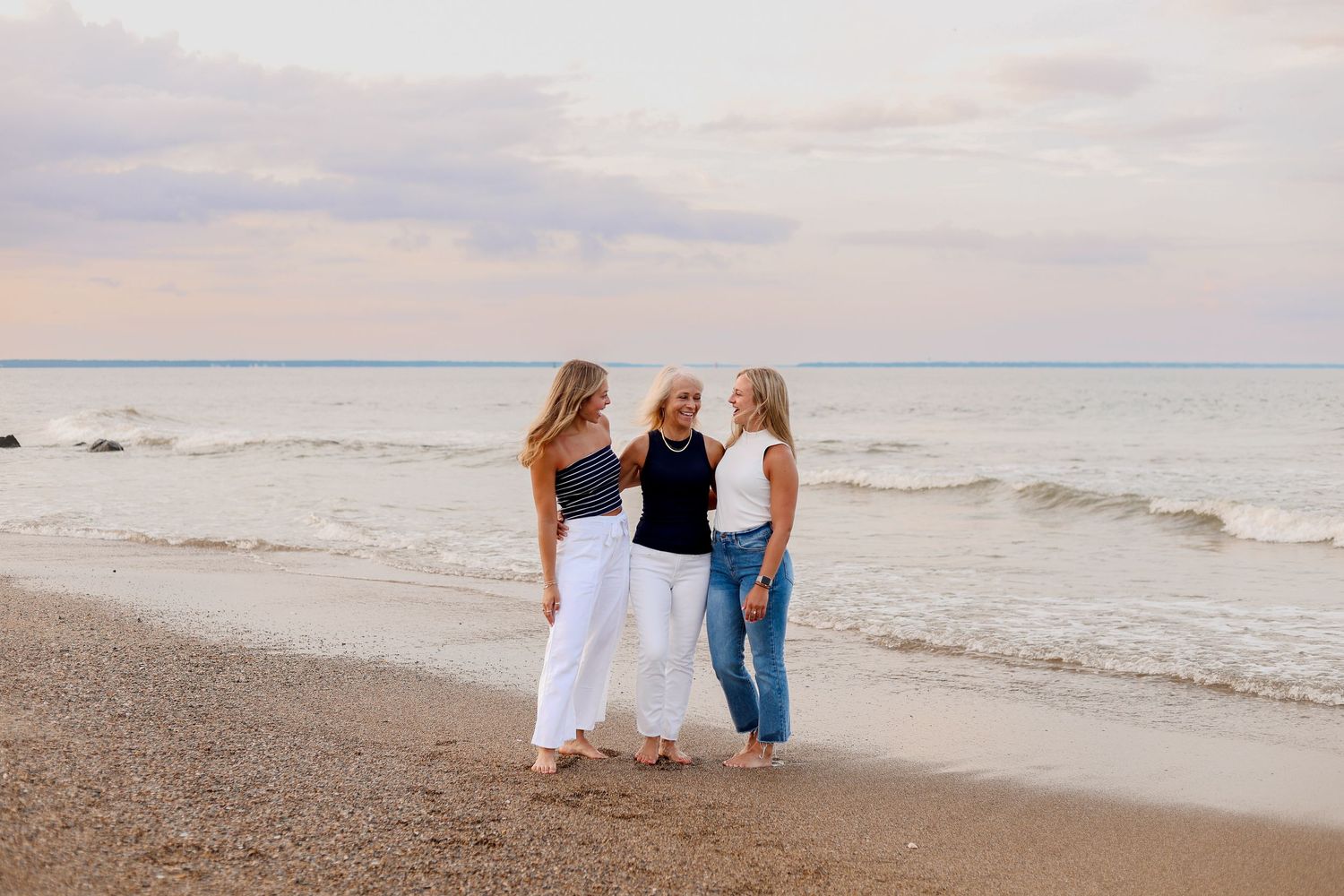 Three friends in casual attire stand together on a beach at sunset with waves rolling in behind them.