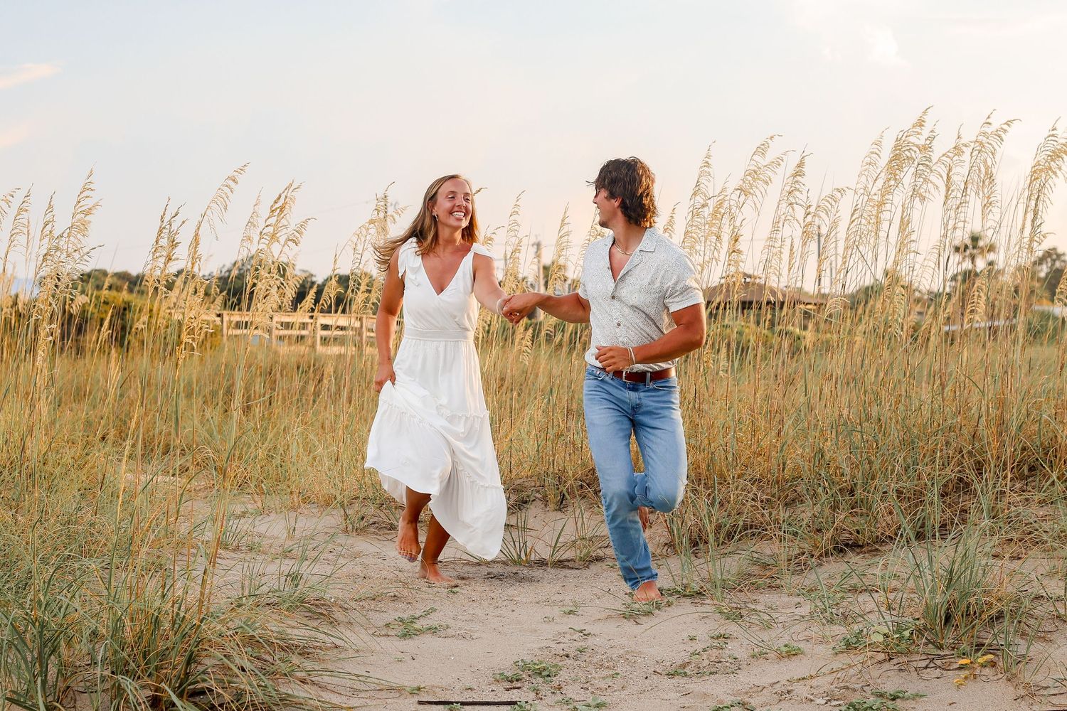 Couple running playfully through beach grass during golden hour.
