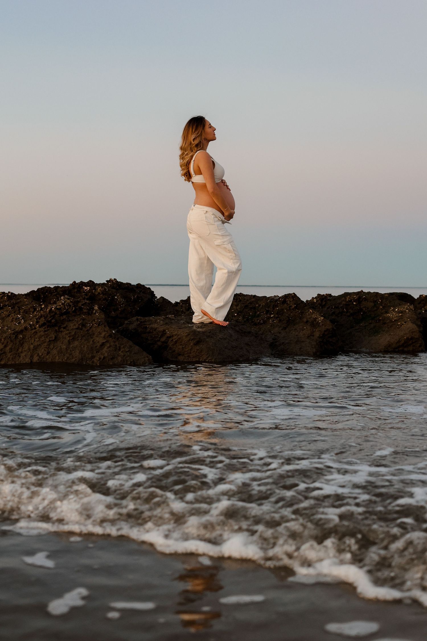 White-clad figure perched on rocky outcrop with ocean waves crashing below at dusk.