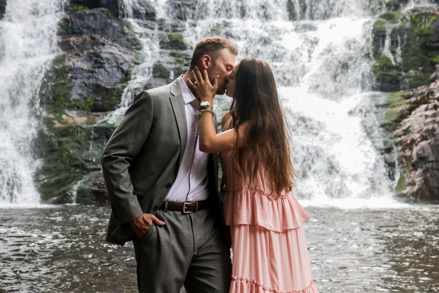 A romantic couple embraces in front of a cascading waterfall wearing a gray suit and pink dress.