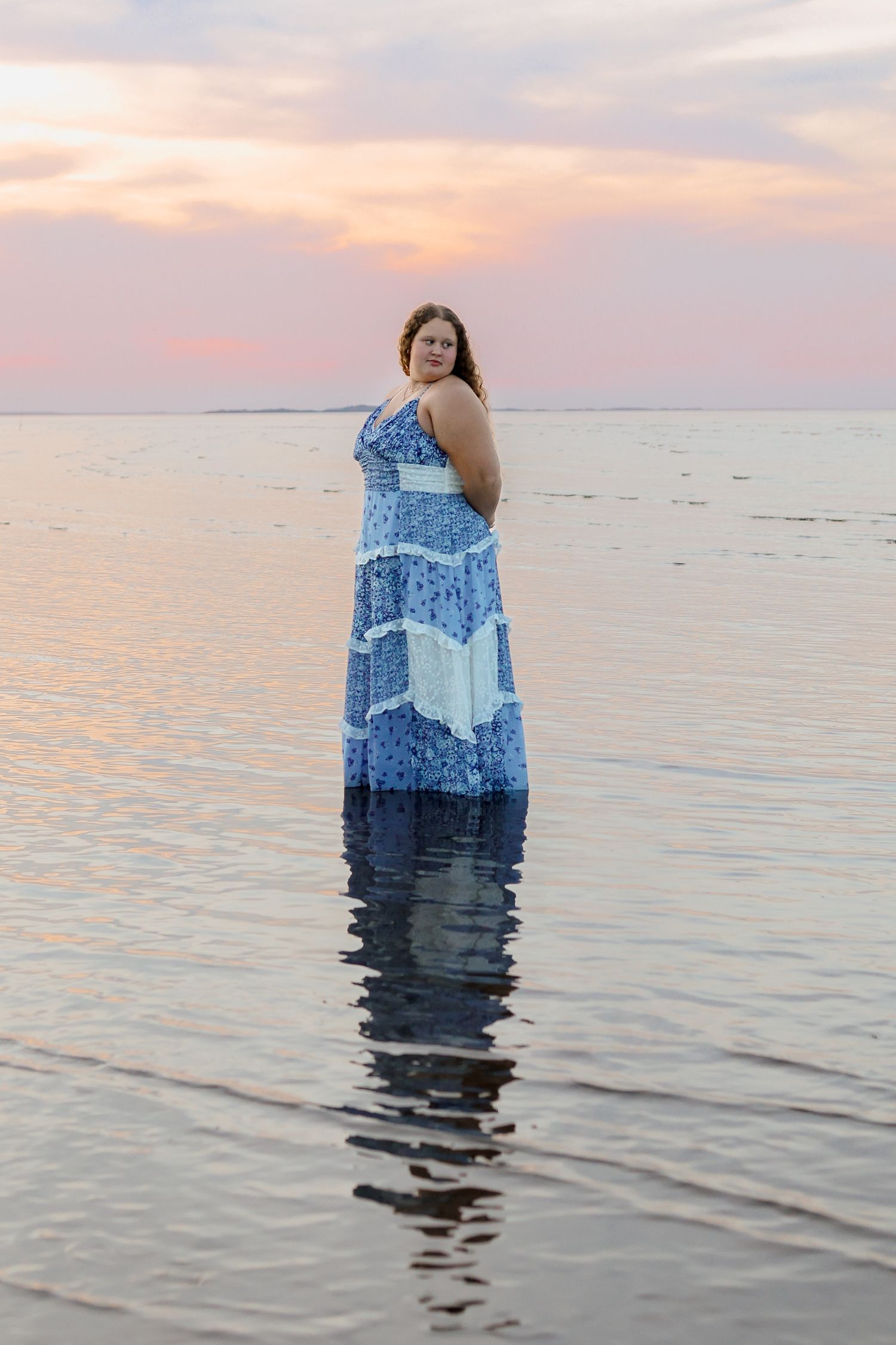 A person wearing a blue patterned maxi dress stands in shallow ocean water during sunset, creating a reflection on the surface.