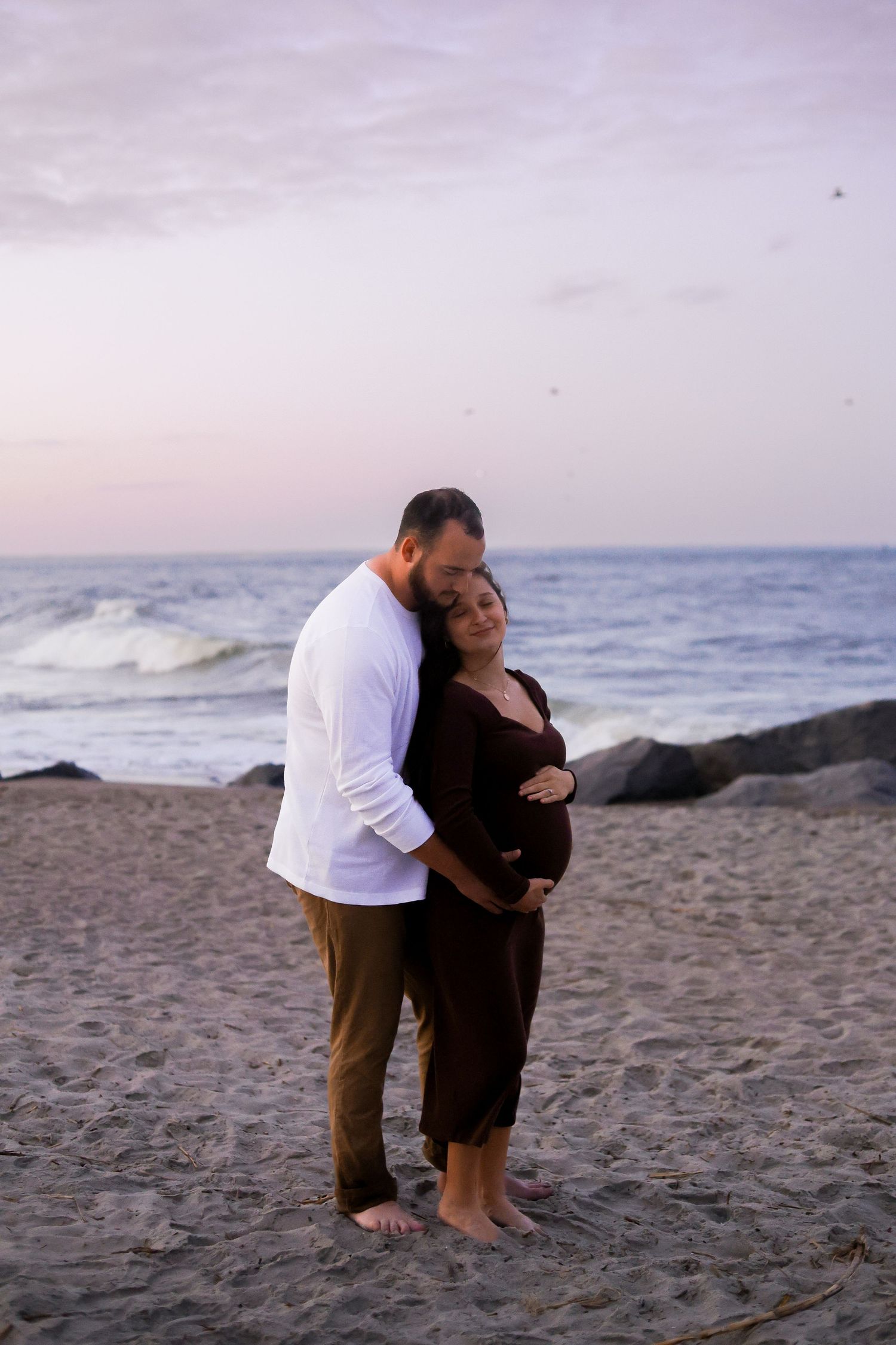 Intimate maternity moment captured on sandy beach at dusk with ocean waves in background.