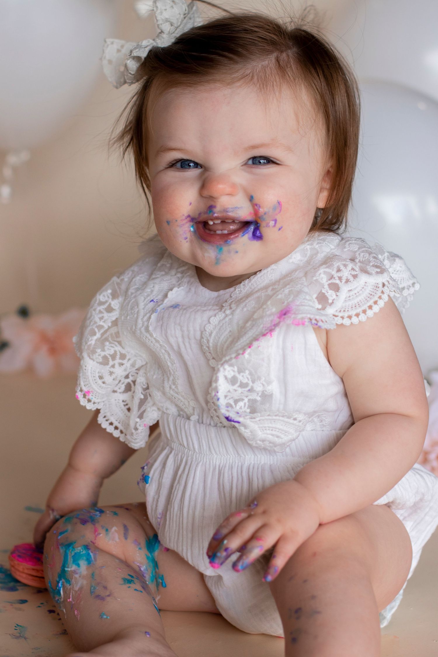 Baby wearing white dress getting messy with colorful cake frosting during cake smash session.