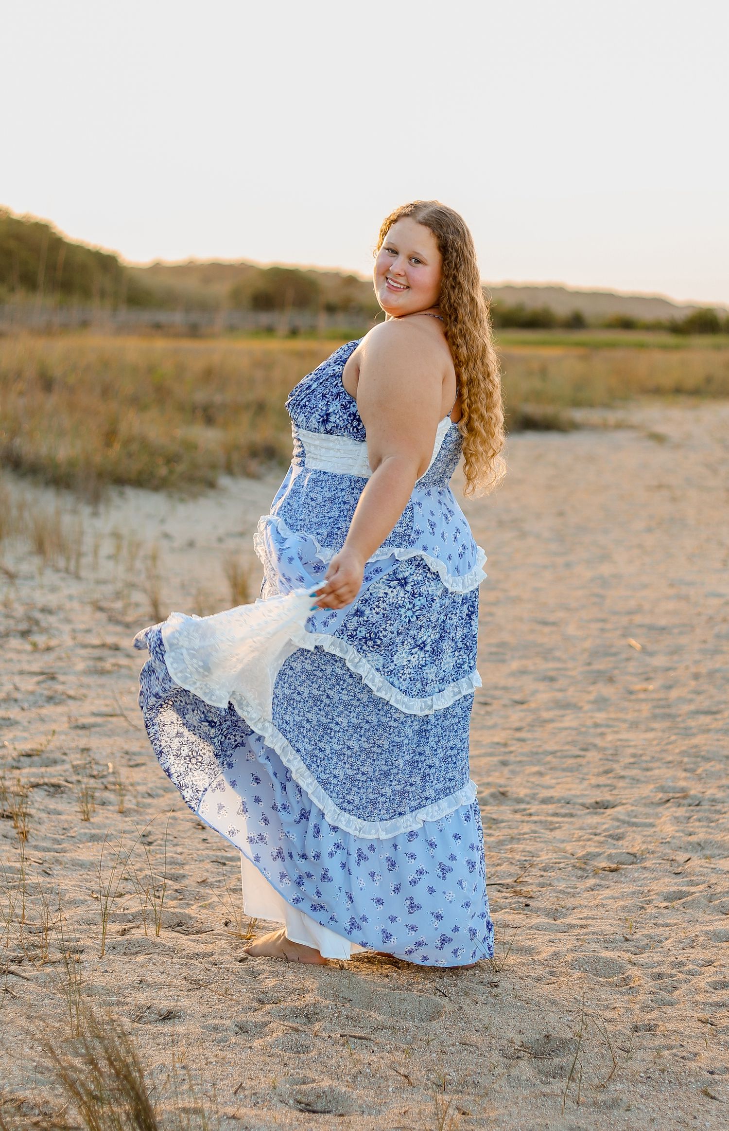 Young woman in a flowing blue and white floral dress twirls playfully on a sandy beach at sunset.