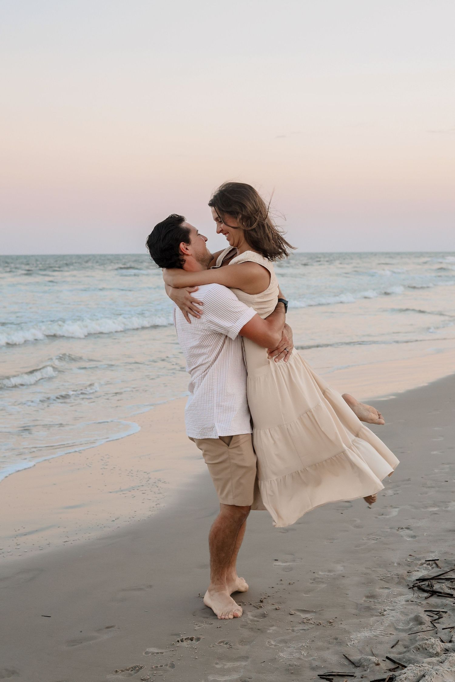A couple embraces and dances together on a sandy beach at sunset with ocean waves in the background.