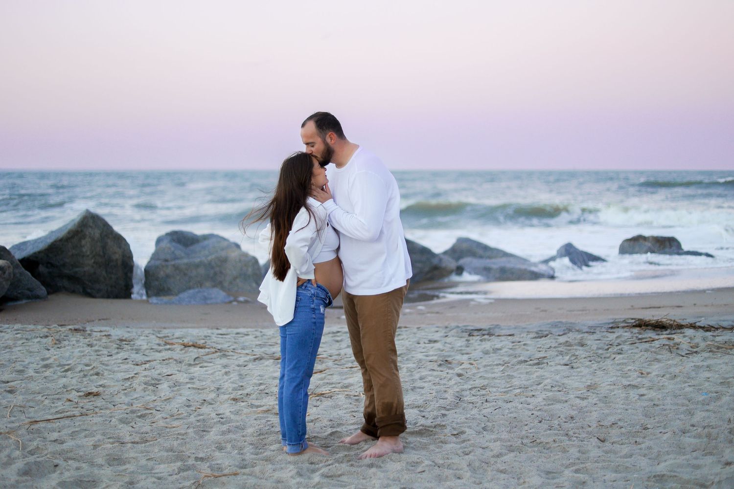 Romantic couple embracing on beach at sunset with rocky shoreline in background.